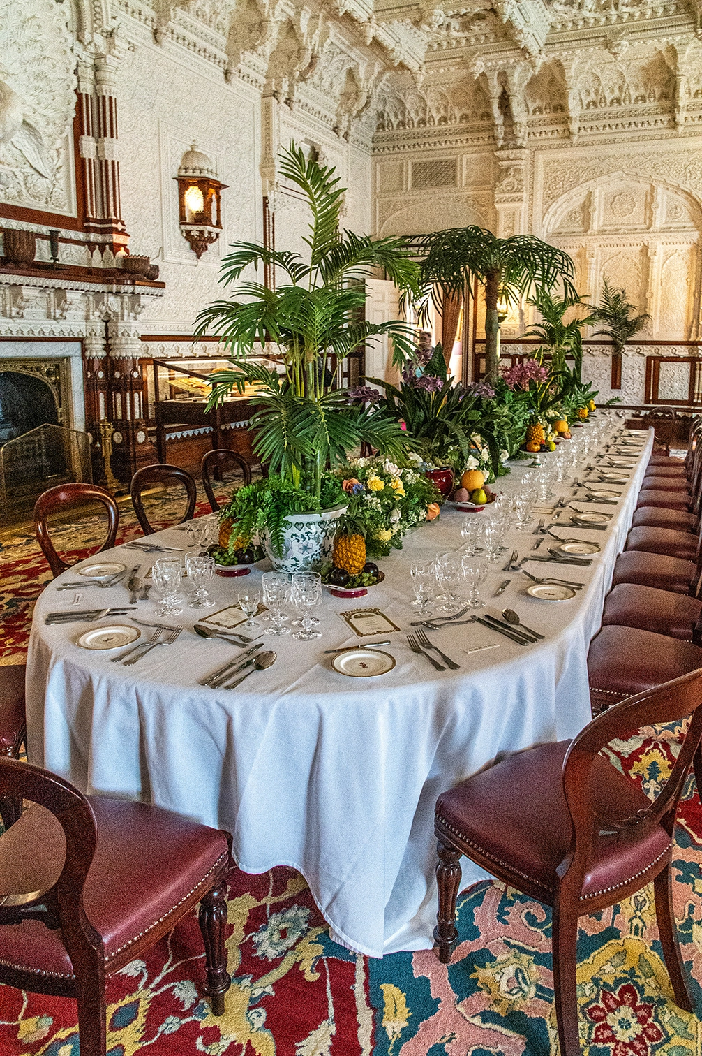 The image depicts an elaborately set dining table in a grand, ornate room. The table is adorned with white tablecloths, fine china, glassware, and cutlery, along with decorative plants and flowers. The room features intricate architectural details, including carved arches and columns, and is furnished with plush, upholstered chairs. The ambiance suggests a formal dining setting, possibly for a banquet or special event.