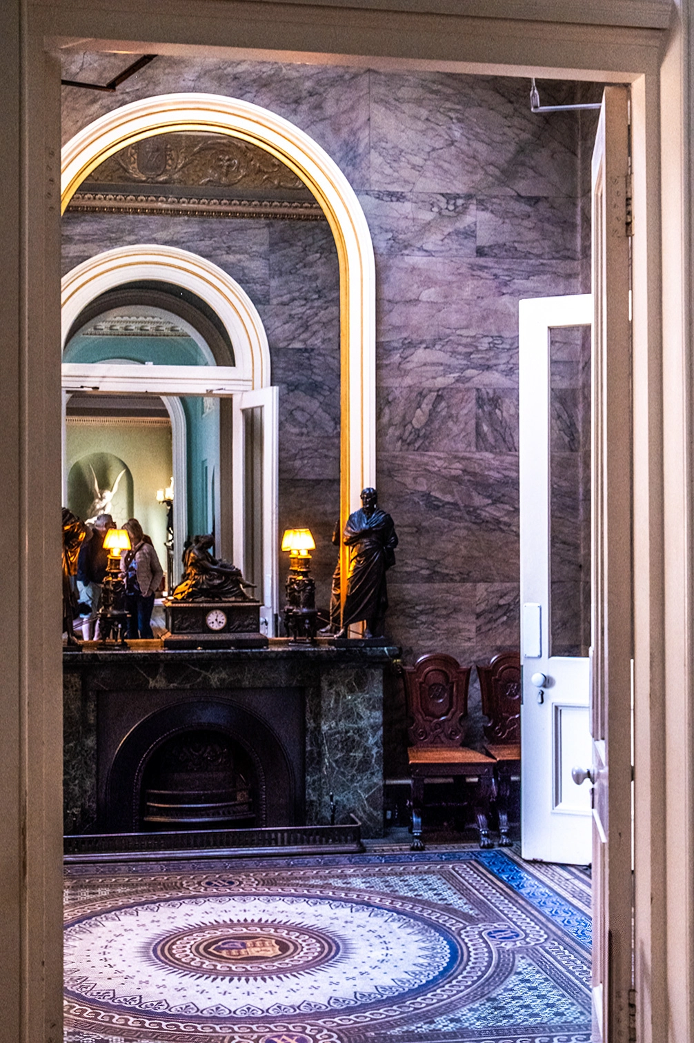 The image depicts an ornate interior space with classical architectural elements. It features a marble fireplace with a clock and statues, intricate floor patterns, and elegant wooden chairs. The room has arched doorways and is adorned with decorative moldings and marble walls. Warm lighting from table lamps adds to the sophisticated ambiance. The space appears to be part of a historical or official building, possibly a museum or government facility.