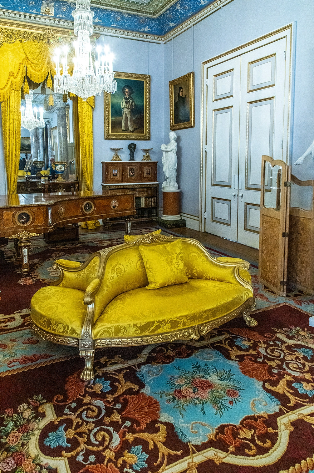 The image depicts an ornate, historical room with a bright yellow chaise lounge as the focal point. The room features intricate decor, including a detailed floral rug, antique wooden furniture, and a crystal chandelier. Paintings and sculptures adorn the walls, and a large mirror is framed with yellow drapes. The overall ambiance suggests a luxurious and well-preserved interior, likely from a palace or a grand estate.