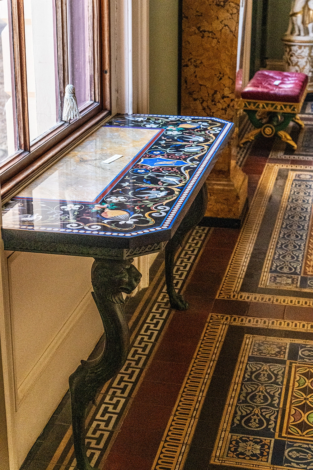 This image showcases a beautifully detailed console table with a decorative inlay on its surface. It's positioned next to a window, with a tasselled curtain tie-back visible. The table's legs are carved into the shape of lion's heads, adding an element of grandeur. The floor features an intricate tiled pattern, and in the background, a glimpse of a red upholstered stool and a decorative column can be seen.