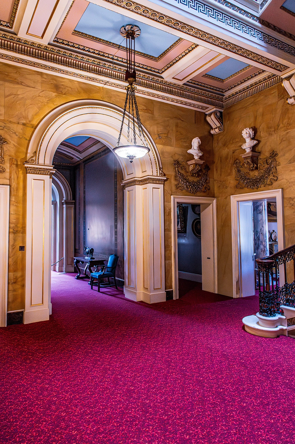 The image depicts an elegant, ornate hallway with high ceilings adorned with intricate moldings and a patterned carpet. The hallway features classical architectural elements such as arches, columns, and decorative busts. A chandelier hangs from the ceiling, and there is a table with a blue chair positioned along the wall. The walls are decorated with marble-like panels and there are several doorways leading to other rooms. The overall ambiance is one of opulence and historical grandeur.
