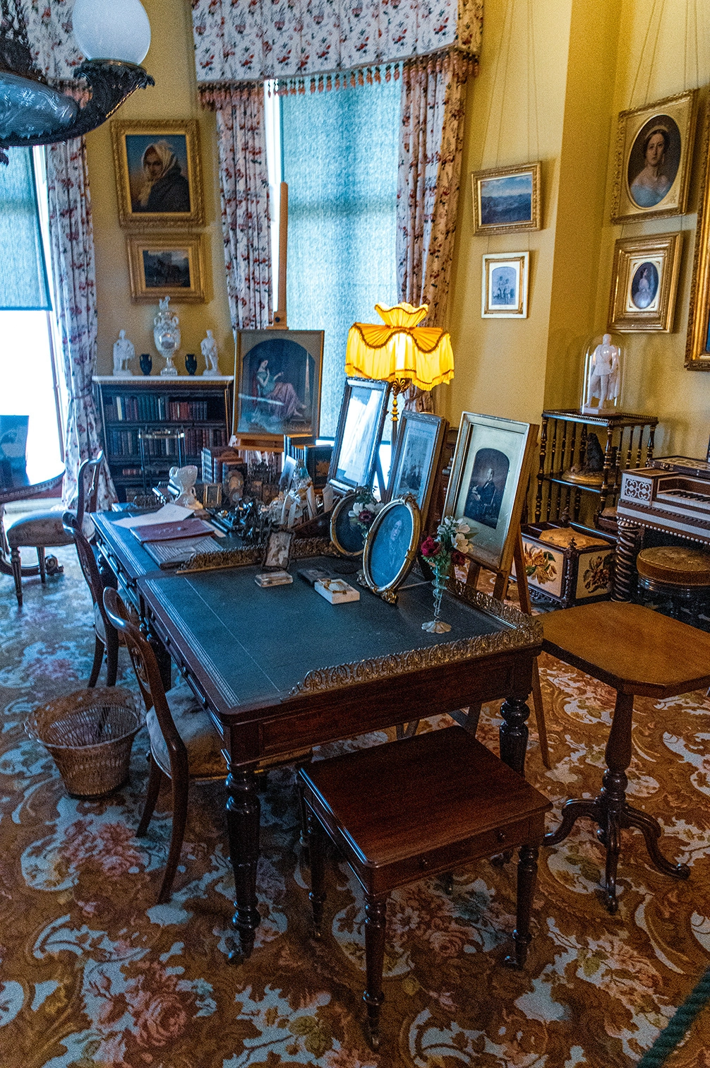 The image depicts a vintage study room adorned with antique furniture and decor. A large wooden desk is central, surrounded by various chairs and a footstool. The desk holds several framed photographs and personal items. The room features ornate curtains, a bookshelf filled with books and sculptures, and multiple framed portraits on the walls. The overall ambiance is one of historical elegance and sophistication.