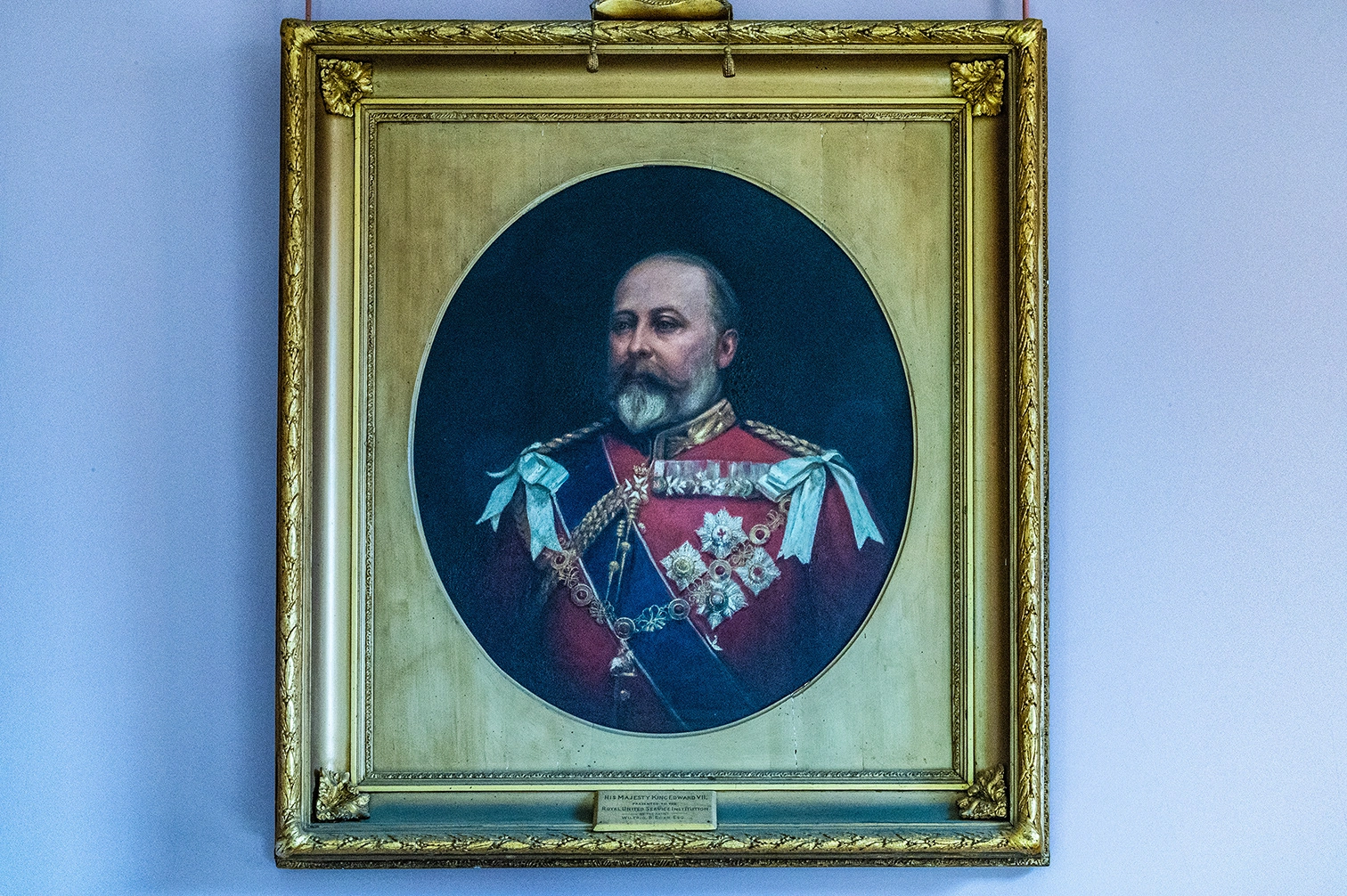 This is a formal portrait of King Edward VII, framed in an ornate gold frame against a light blue wall. The painting itself is circular and depicts the King from the chest up, wearing a striking red military uniform adorned with a blue sash and numerous medals. He has a distinguished grey beard and moustache, and a serious expression. 