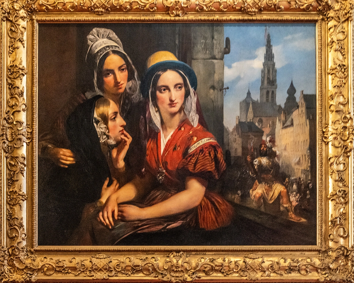 The image depicts a framed painting with three women in traditional attire prominently featured in the foreground. The woman in the center is wearing a red dress and a blue headscarf, while the other two women are dressed in darker clothing with white head coverings. The background shows a historical cityscape with a tall spire and other architectural elements, suggesting a European setting. The ornate golden frame adds a decorative touch to the overall presentation.