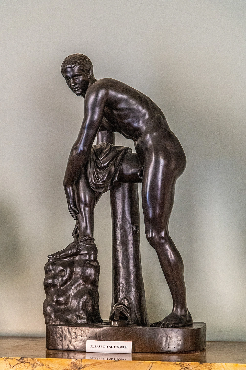 The image depicts a bronze statue of a young male figure in a dynamic pose, leaning forward with one foot on a rock-like structure. The statue is positioned on a marble base, and there is a sign in front of it that reads 'PLEASE DO NOT TOUCH.' The figure appears to be in a state of motion or exertion, with detailed musculature and a draped cloth partially covering his lower body.