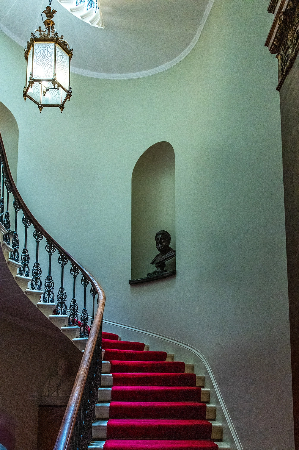The image depicts an elegant interior staircase with a red carpet runner. The staircase features a wooden handrail and ornate wrought iron balusters. A decorative lantern hangs from the ceiling, illuminating the area. The walls are painted a light green color, and there is a small alcove with a bust sculpture. The overall aesthetic is classic and sophisticated.