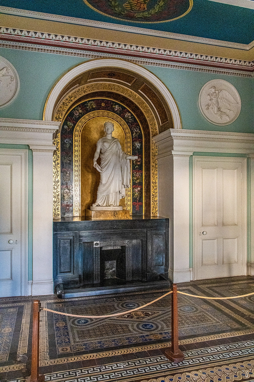 The image depicts an ornate interior room with classical architectural elements. Central to the scene is a statue of a robed figure set within an arched alcove adorned with intricate gold and floral designs. The room features a green and gold color scheme, with detailed moldings and decorative motifs. A fireplace is situated below the statue, and the floor is covered with a richly patterned carpet. The area is cordoned off, suggesting it may be part of a historical or museum exhibit.