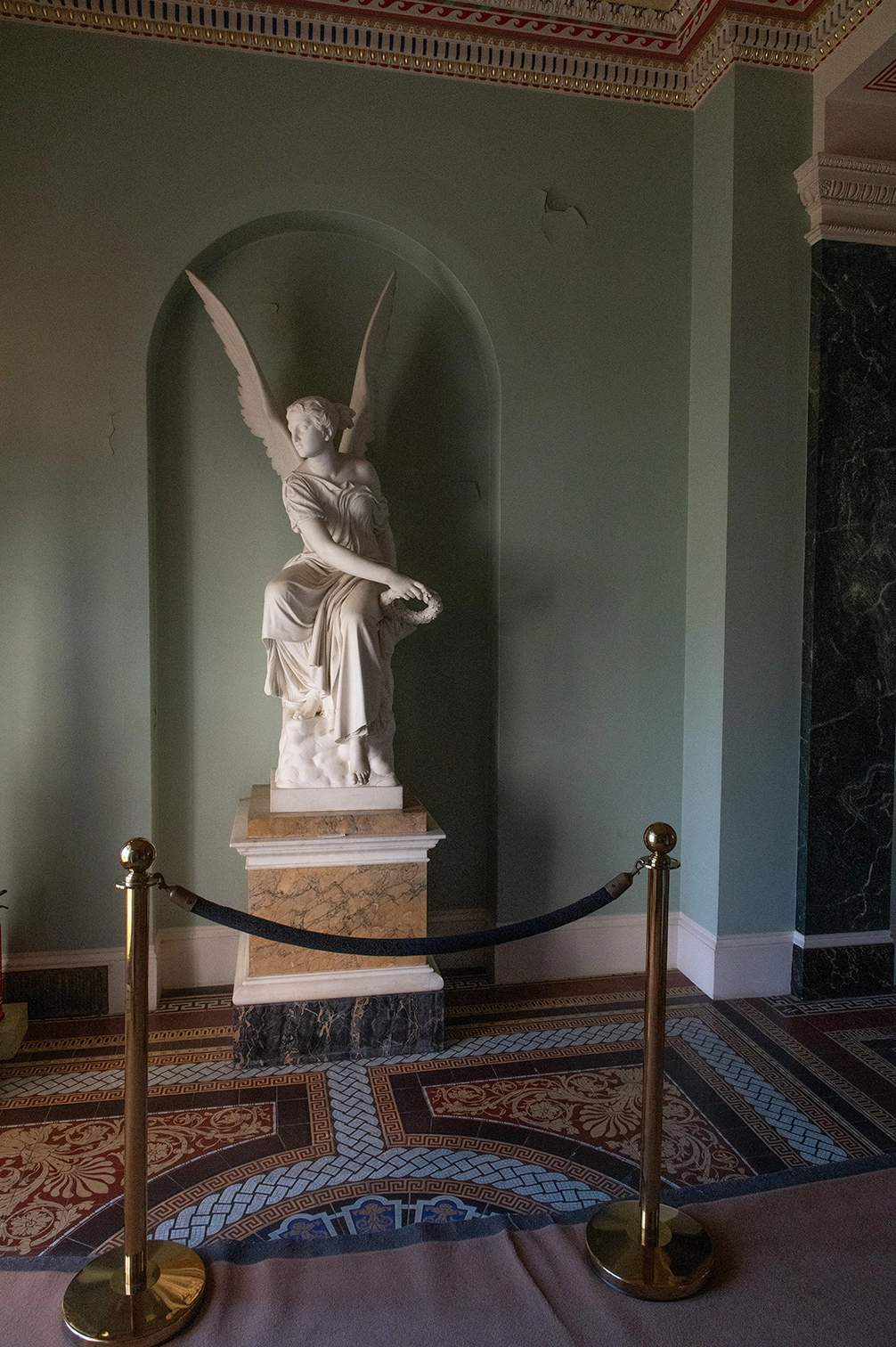 The image depicts an indoor scene featuring a classical statue of a winged figure, likely an angel, placed within an arched alcove. The statue is positioned on a pedestal and is cordoned off by a black rope supported by two golden stanchions. The room has ornate architectural details, including decorative molding on the ceiling and walls, and a patterned floor with intricate designs. The overall ambiance suggests a historical or culturally significant setting, possibly a museum or a grand estate.