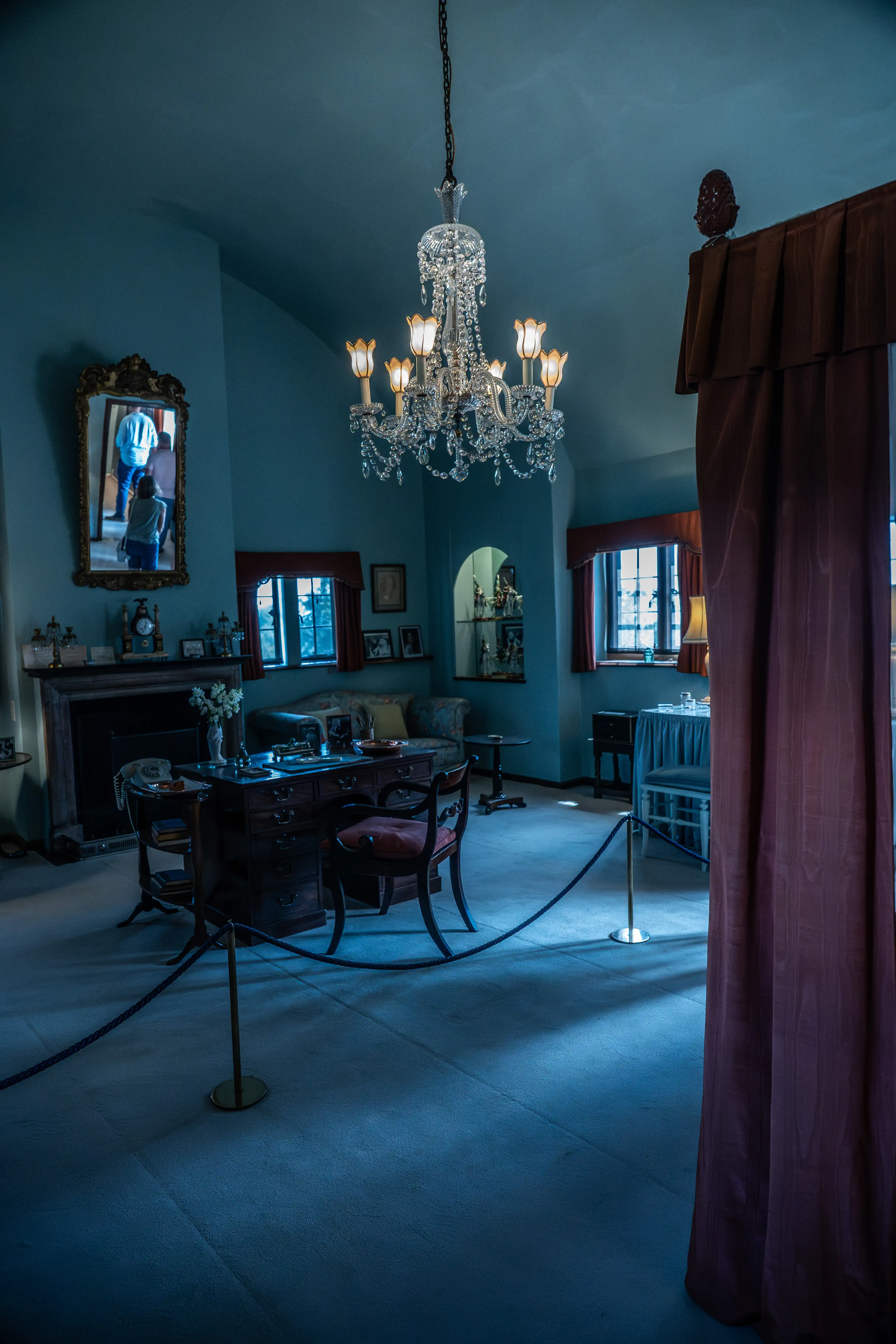 The image depicts a vintage-style room with a chandelier hanging from the ceiling. The room features a desk with a chair, a fireplace with various decorative items, and a seating area with a couch and tables. There is a large mirror above the fireplace and several framed pictures on the walls. The room is cordoned off, suggesting it might be part of a museum or historical site.