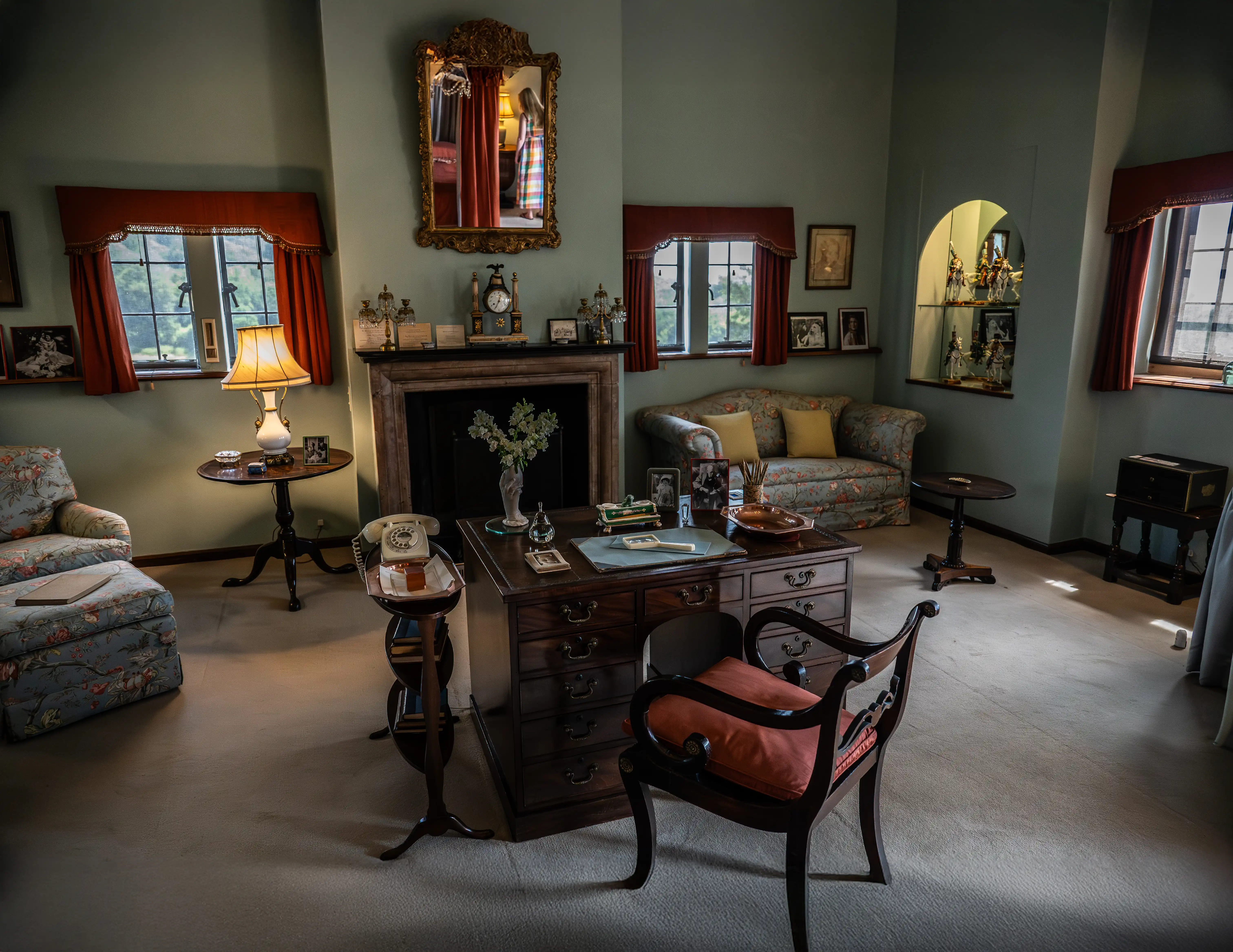 The image depicts a well-furnished, vintage-style living room with floral-patterned sofas and curtains. The room features a desk with a chair, a fireplace adorned with various ornaments, and several framed photographs. There are also multiple tables with lamps, a telephone, and various decorative items. The room is bathed in soft, natural light coming through the windows, creating a cozy and inviting atmosphere.