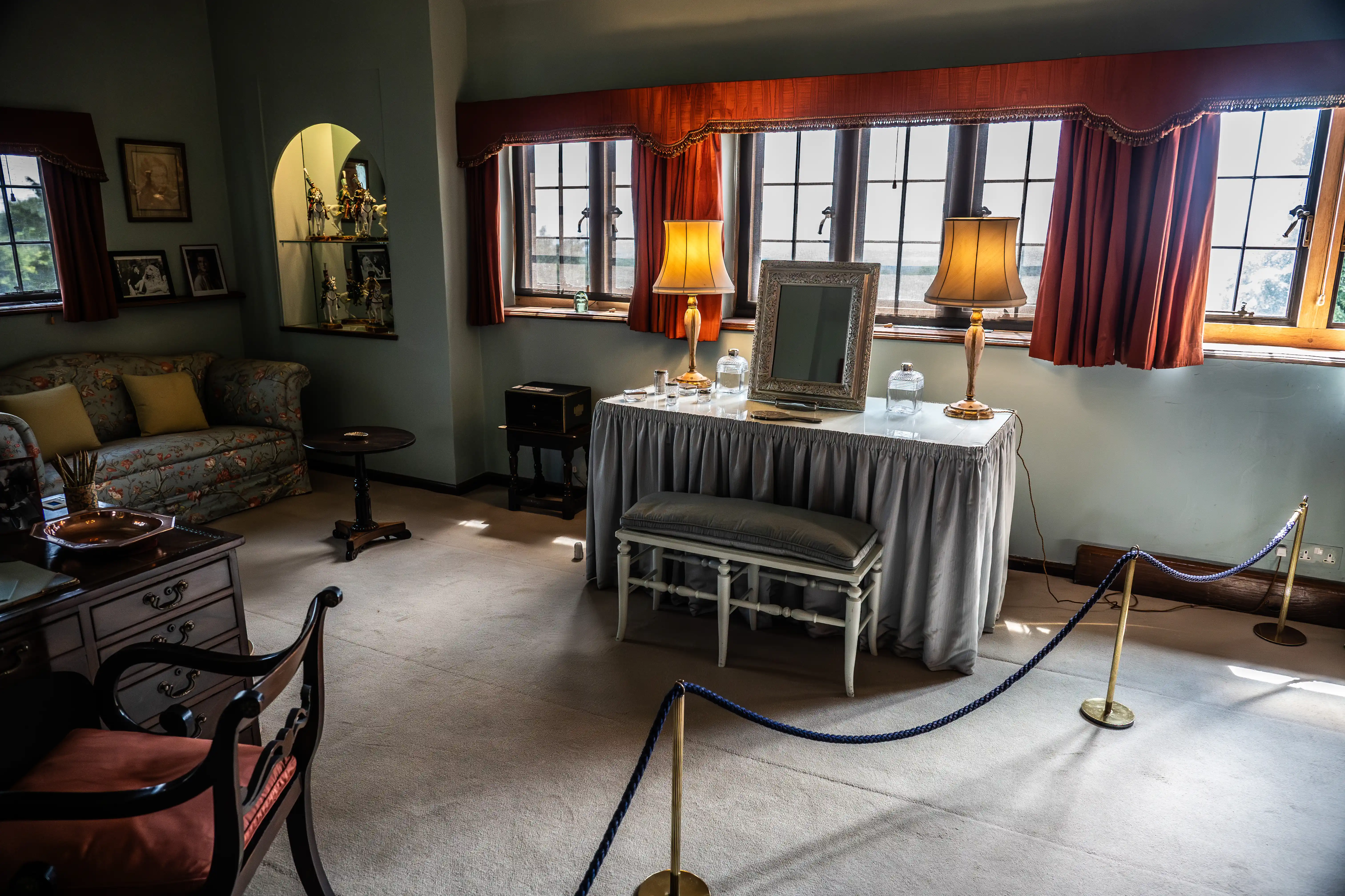 The image depicts a well-furnished, vintage-style room with a floral sofa, a vanity table with a mirror and two lamps, and a small seating area with a table and chairs. The room is cordoned off with a rope barrier, suggesting it might be part of a museum or historical site. The decor includes framed photographs, an ornate shelf with figurines, and large windows adorned with red curtains, allowing natural light to fill the space.
