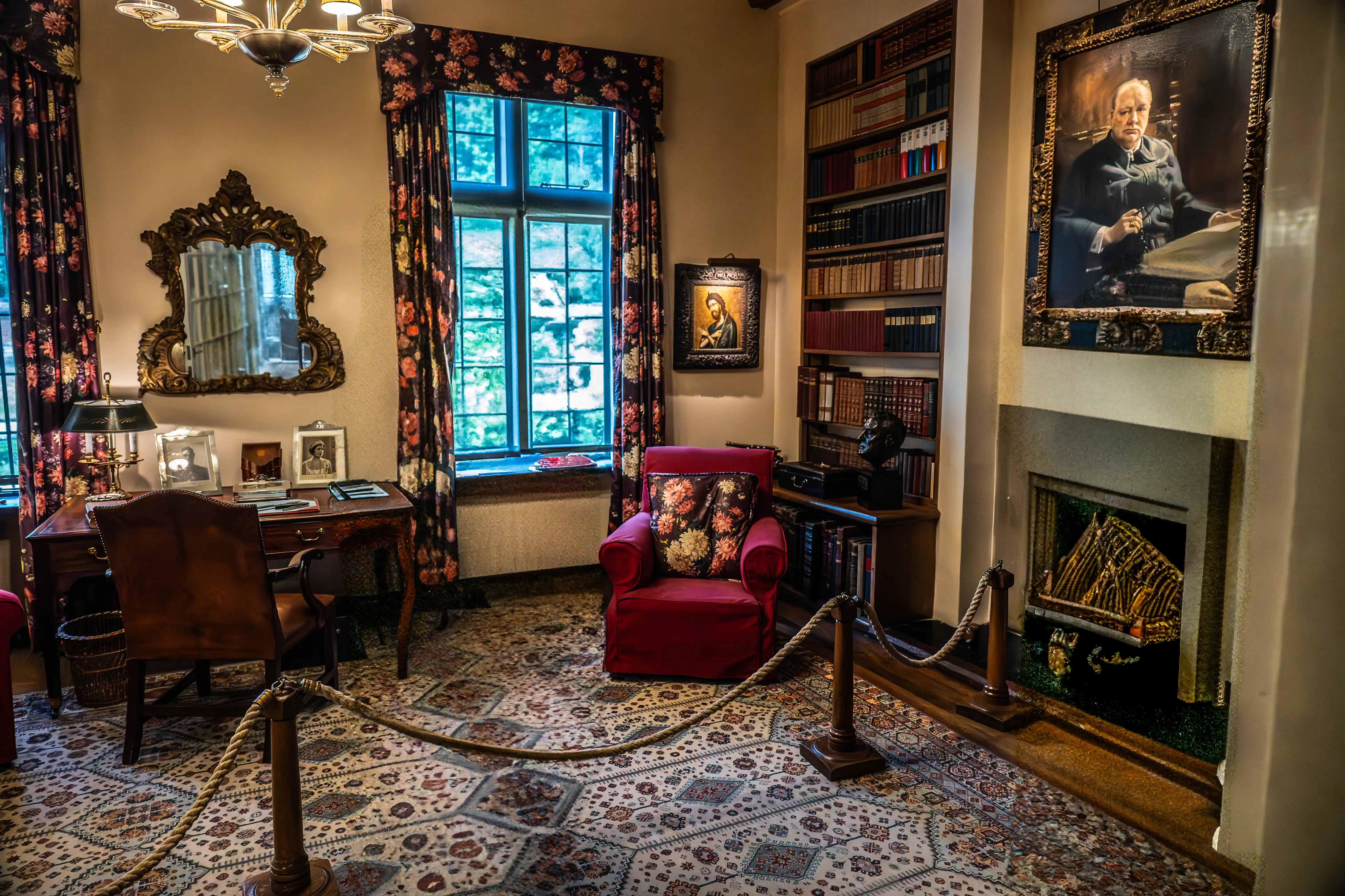 The image depicts a well-furnished, classic study room. Key features include a wooden desk with a chair, a red armchair, a bookshelf filled with books, a fireplace, and ornate decorations such as a large mirror and framed portraits. The room is cordoned off with a rope barrier, suggesting it may be part of a museum or historical site. The curtains, rug, and overall decor contribute to a vintage aesthetic.