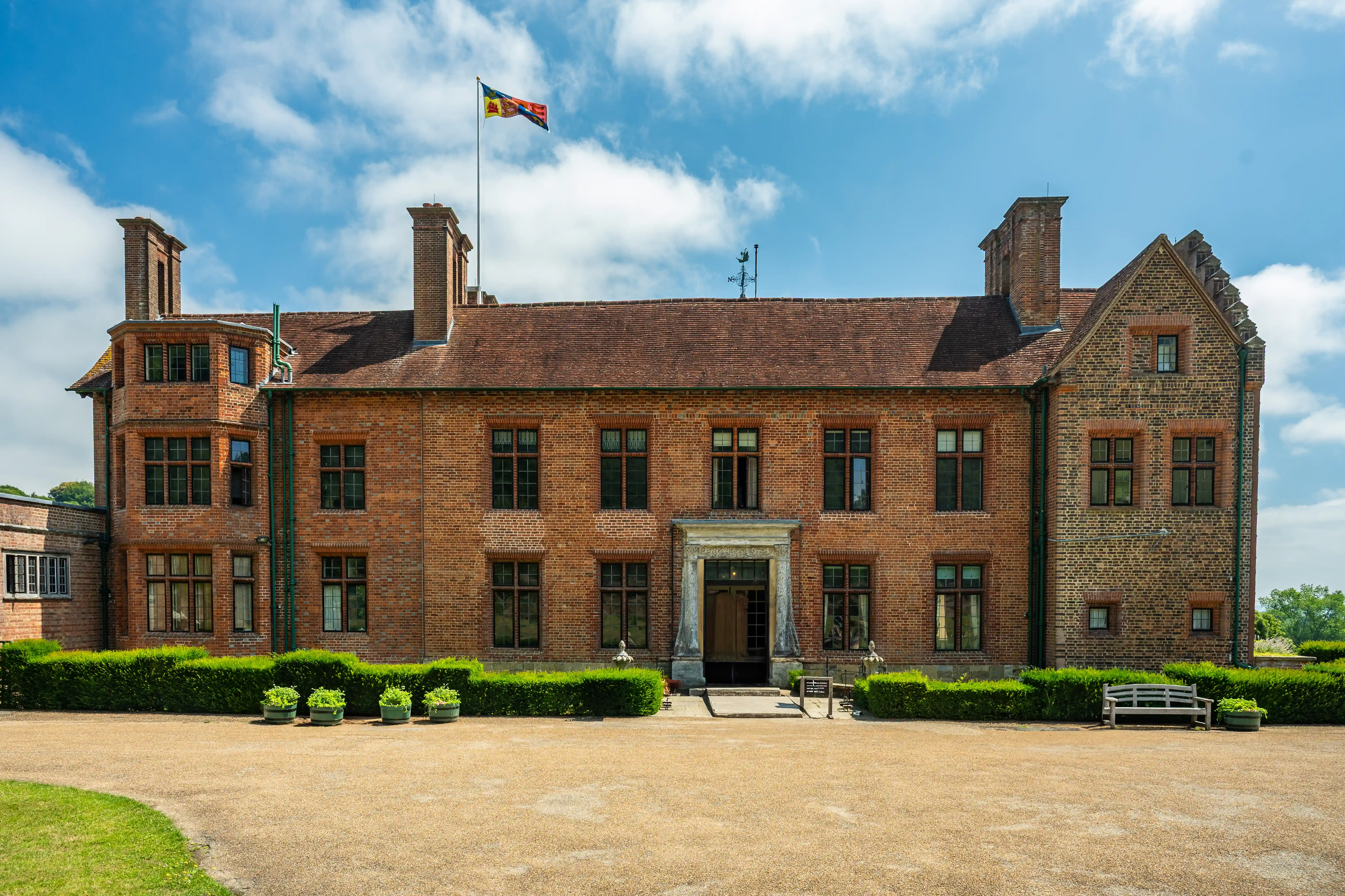 The image depicts a large, historic brick mansion with a symmetrical facade, multiple chimneys, and a central entrance. The building has two main wings and a flag flying atop the roof. The front is adorned with well-maintained shrubs and potted plants, and there is a gravel driveway leading up to the entrance. The sky is clear with some scattered clouds, suggesting a bright and sunny day.