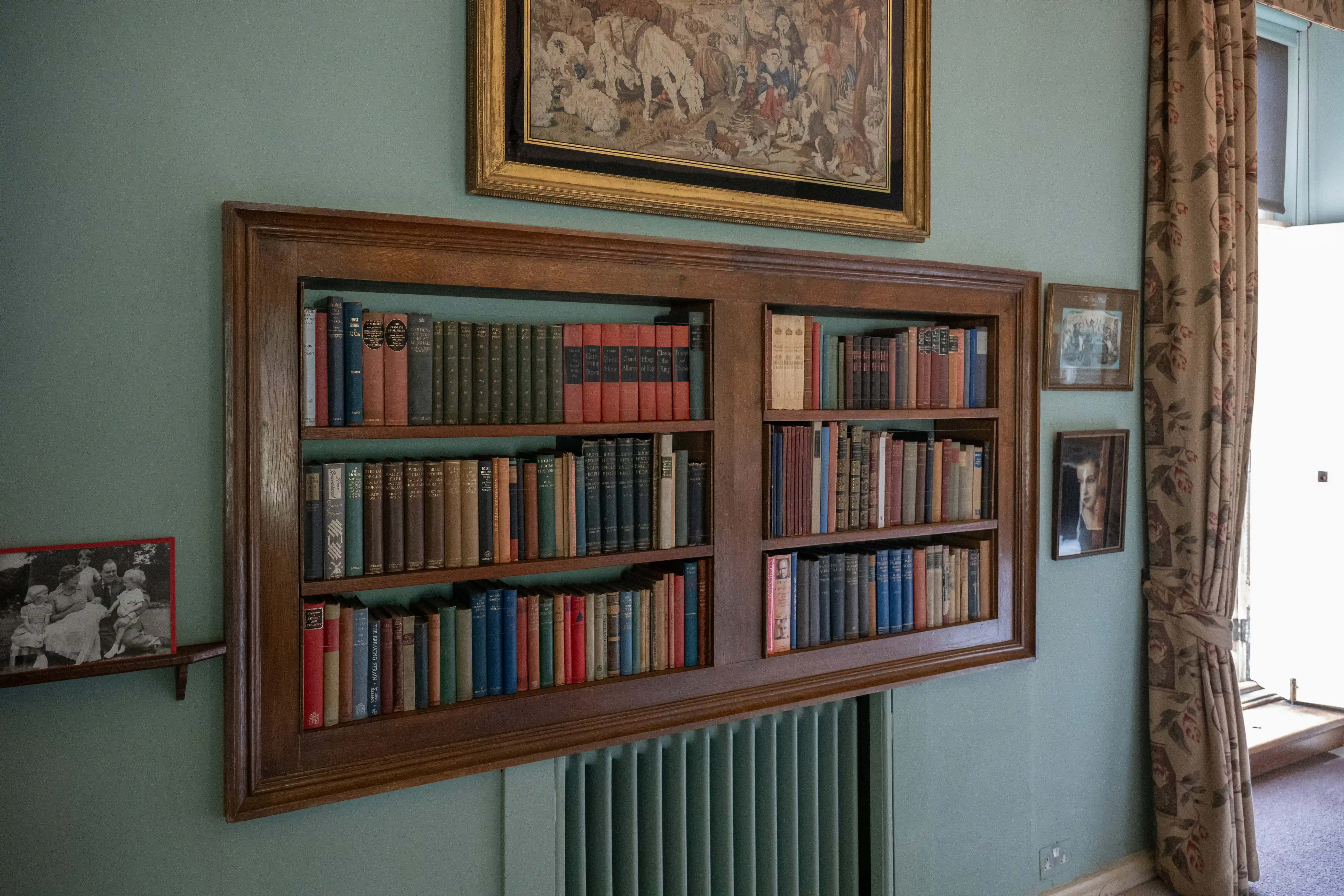 The image depicts a built-in wooden bookshelf filled with a variety of books, organized neatly on the shelves. Above the bookshelf, there is a framed tapestry or artwork. To the right of the bookshelf, there are framed photographs, and a curtain partially covers a window or doorway. The room has a vintage or classic decor style, with light green walls and a radiator below the bookshelf.