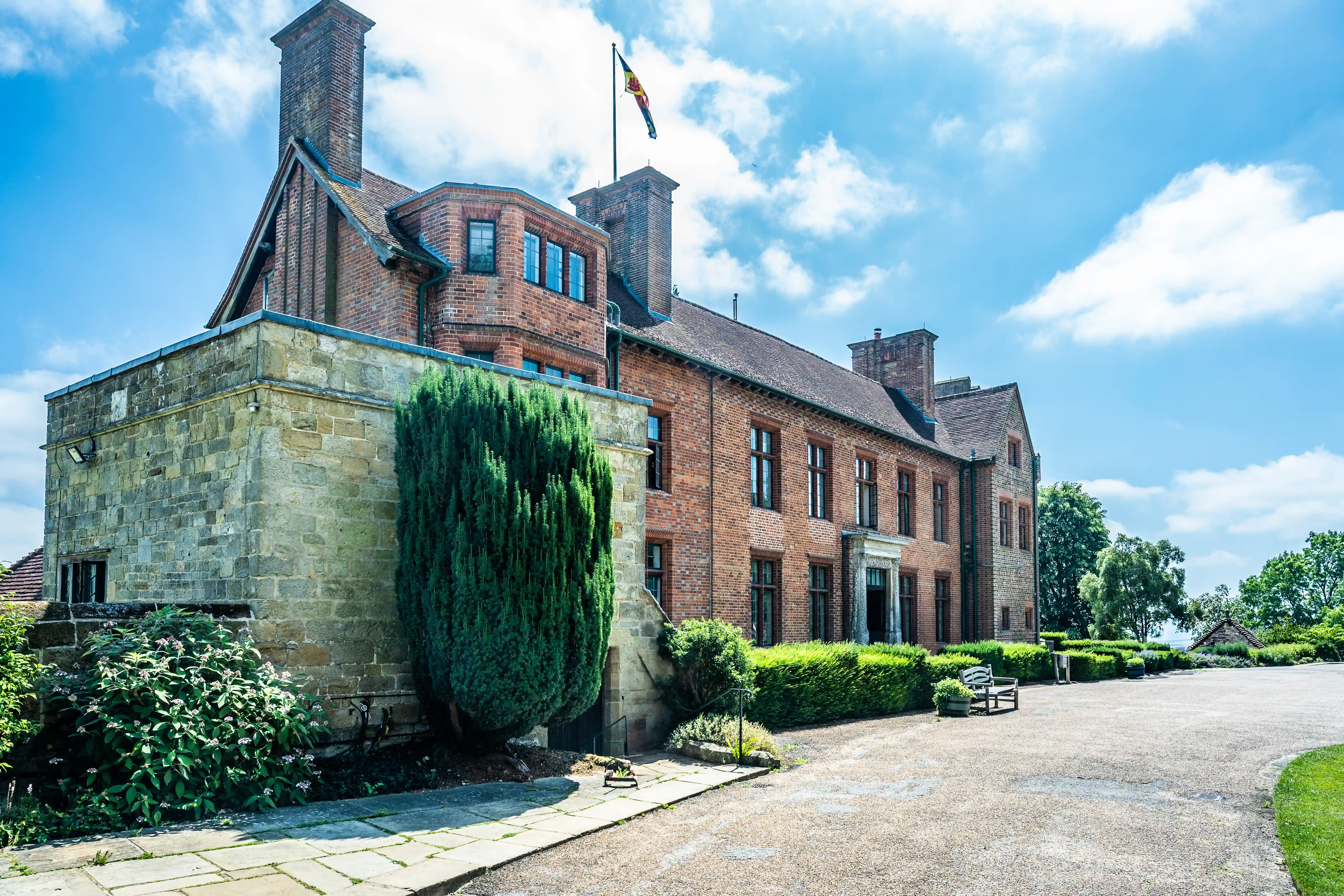 The image depicts a large, historic building constructed primarily of brick and stone. The architecture features multiple chimneys, large windows, and a prominent stone section on the left side. The building is surrounded by well-maintained gardens with various plants and trees, and a flag is visible on top of the structure. The sky is clear with some scattered clouds, indicating a bright and sunny day.