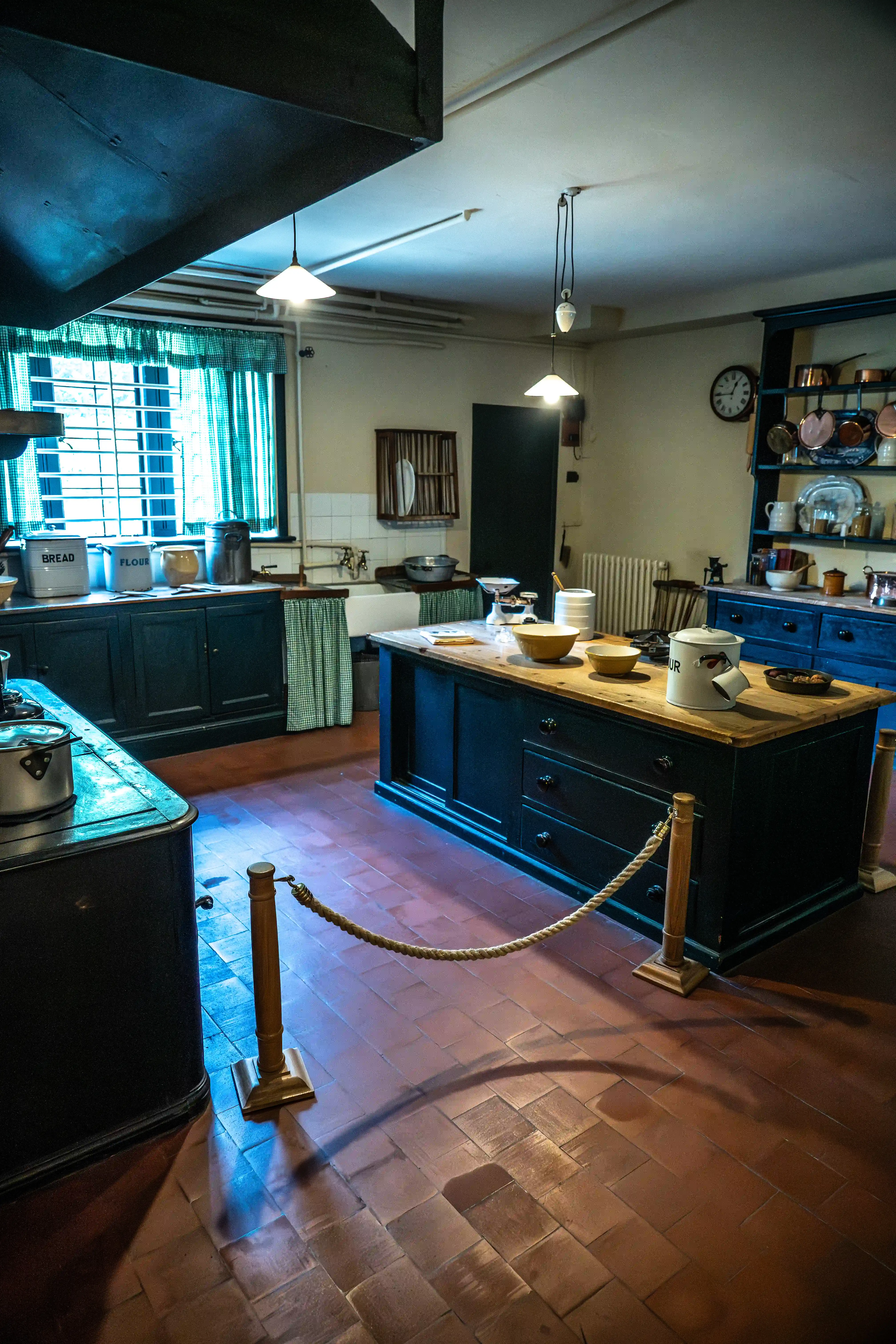 The image depicts a traditional kitchen with a rustic design, featuring dark blue cabinets, a central island with a wooden countertop, and various kitchen utensils and containers. The floor is tiled with reddish-brown tiles, and there are two pendant lights hanging from the ceiling. A rope barrier is present, suggesting that this kitchen might be part of a museum or historical exhibit. The overall ambiance is vintage, with a mix of practical and decorative elements.