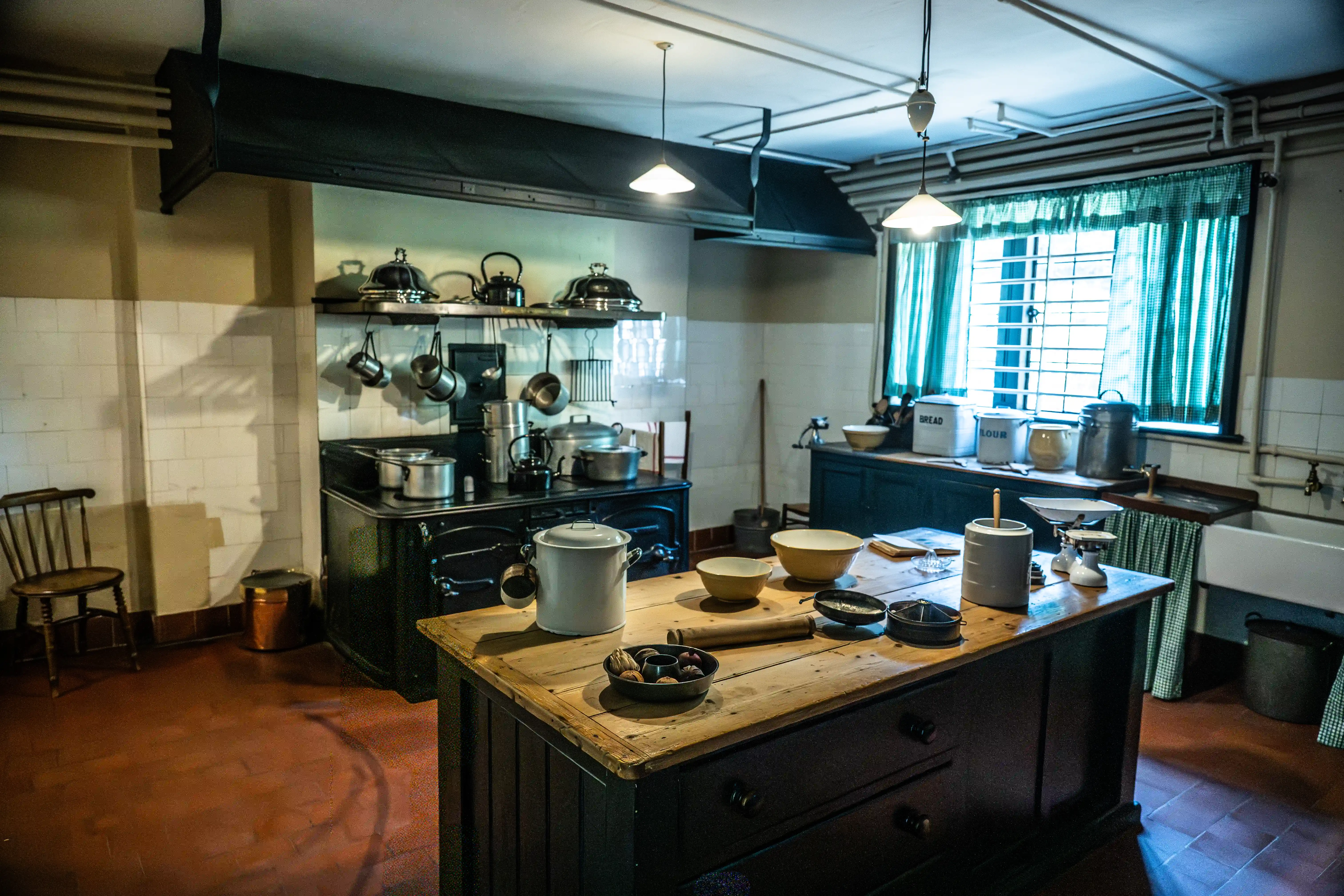 The image depicts a vintage kitchen setup with various traditional kitchen utensils and appliances. The kitchen features a central wooden table with bowls, pots, and other items, surrounded by an old-fashioned stove with hanging pots and pans. There are tiled walls, a window with green curtains, and a few chairs, giving it a historical and rustic appearance. The overall ambiance suggests a well-preserved or recreated kitchen from an earlier era.