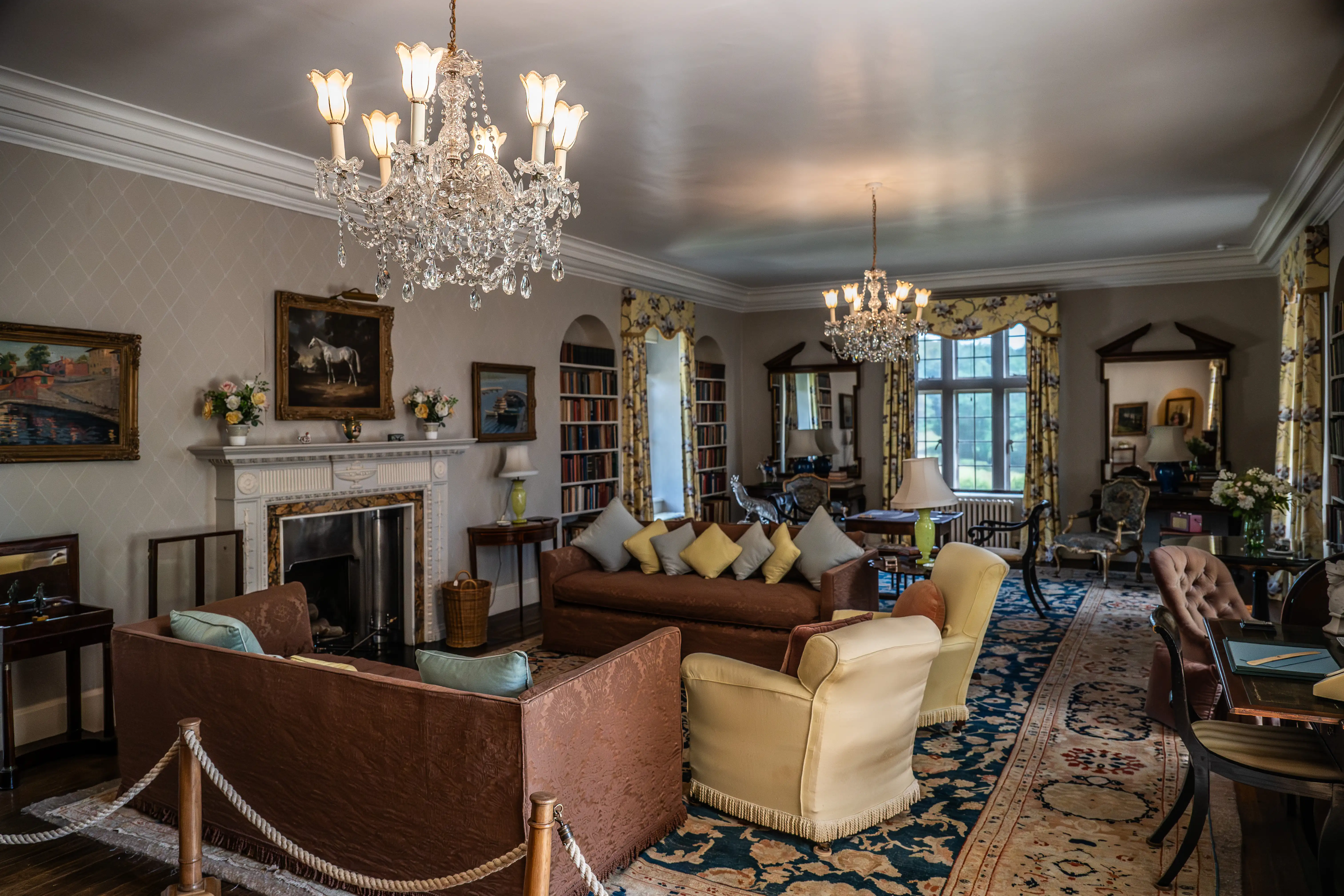 The image depicts an elegantly decorated living room with classic and luxurious furnishings. It features a large, ornate chandelier, plush sofas and armchairs, a fireplace, and numerous decorative items including paintings and vases. The room is well-lit with natural light coming through large windows adorned with elaborate curtains. Bookshelves filled with books line the walls, contributing to a cozy and intellectual atmosphere.