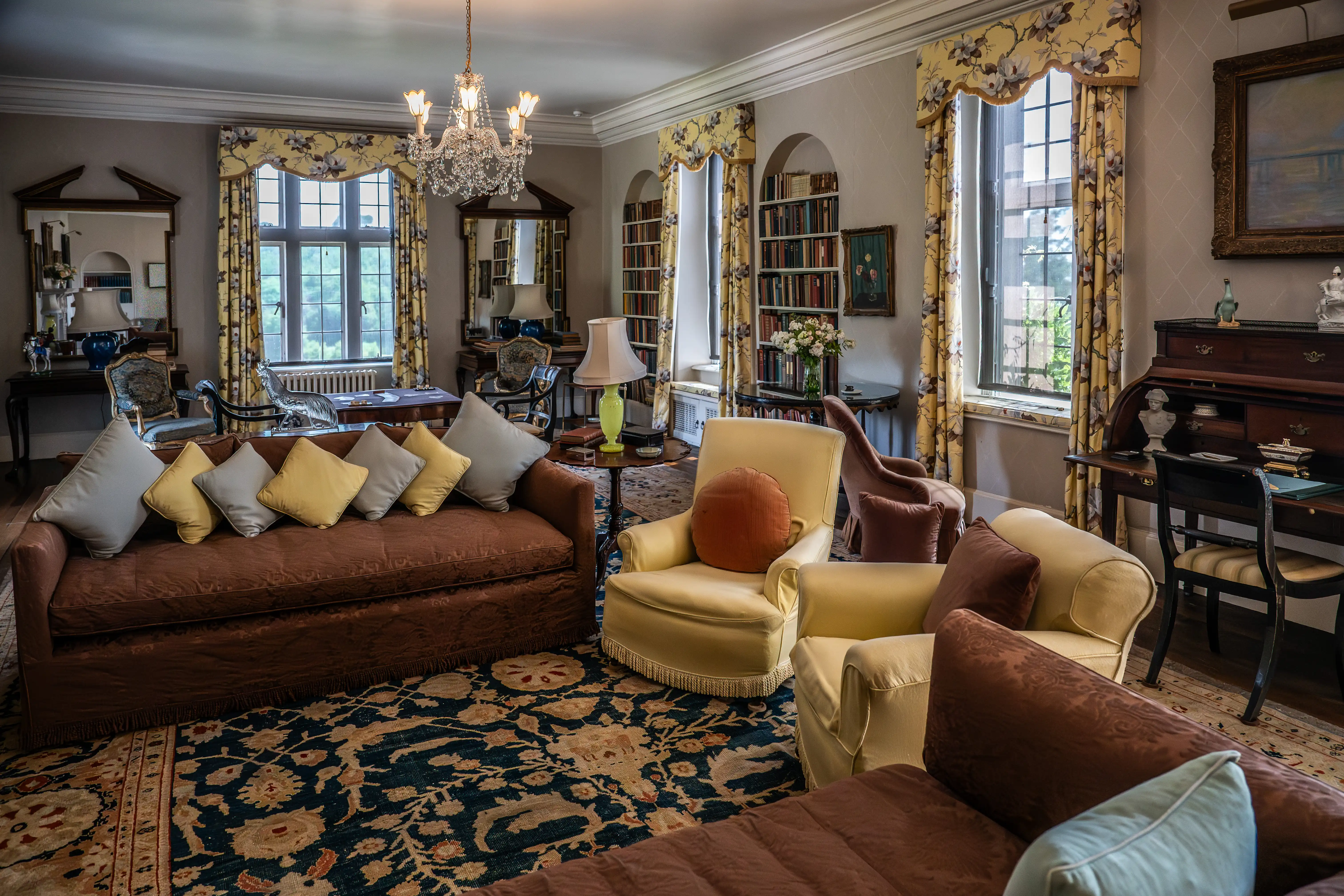 The image depicts a classic and elegantly furnished living room. The room features a brown sofa adorned with multiple cushions, two yellow armchairs, and a patterned rug. There are large windows with floral curtains, a chandelier hanging from the ceiling, and bookshelves filled with books. The decor includes various pieces of furniture and decorative items, creating a cozy and sophisticated atmosphere.