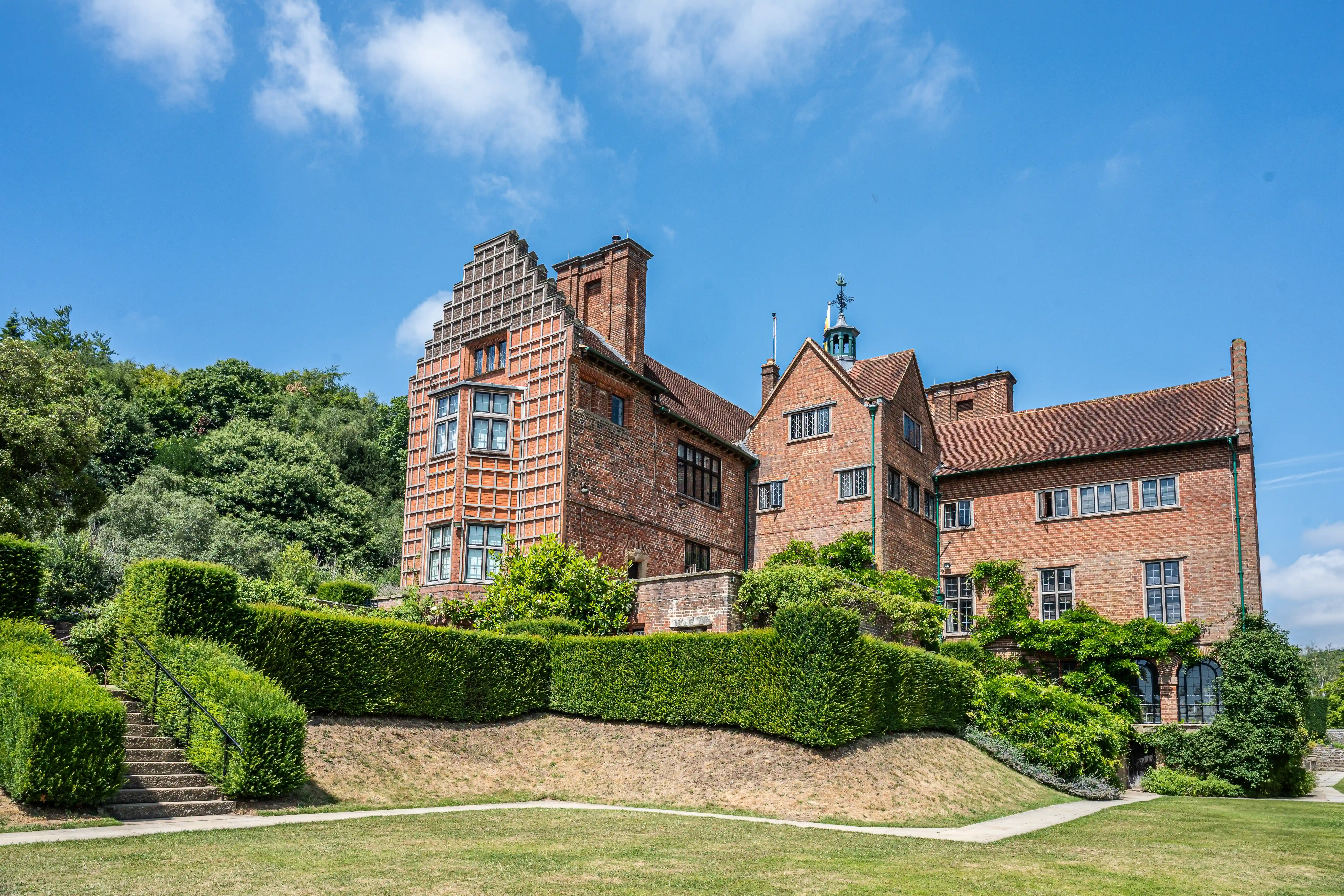 The image depicts a large, historic brick building with distinct architectural features, including a stepped gable and multiple chimneys. The structure is surrounded by well-maintained greenery and hedges, with a set of stairs leading up to the building. The sky is clear and blue, suggesting a bright and sunny day. The overall scene conveys a sense of grandeur and tranquility.