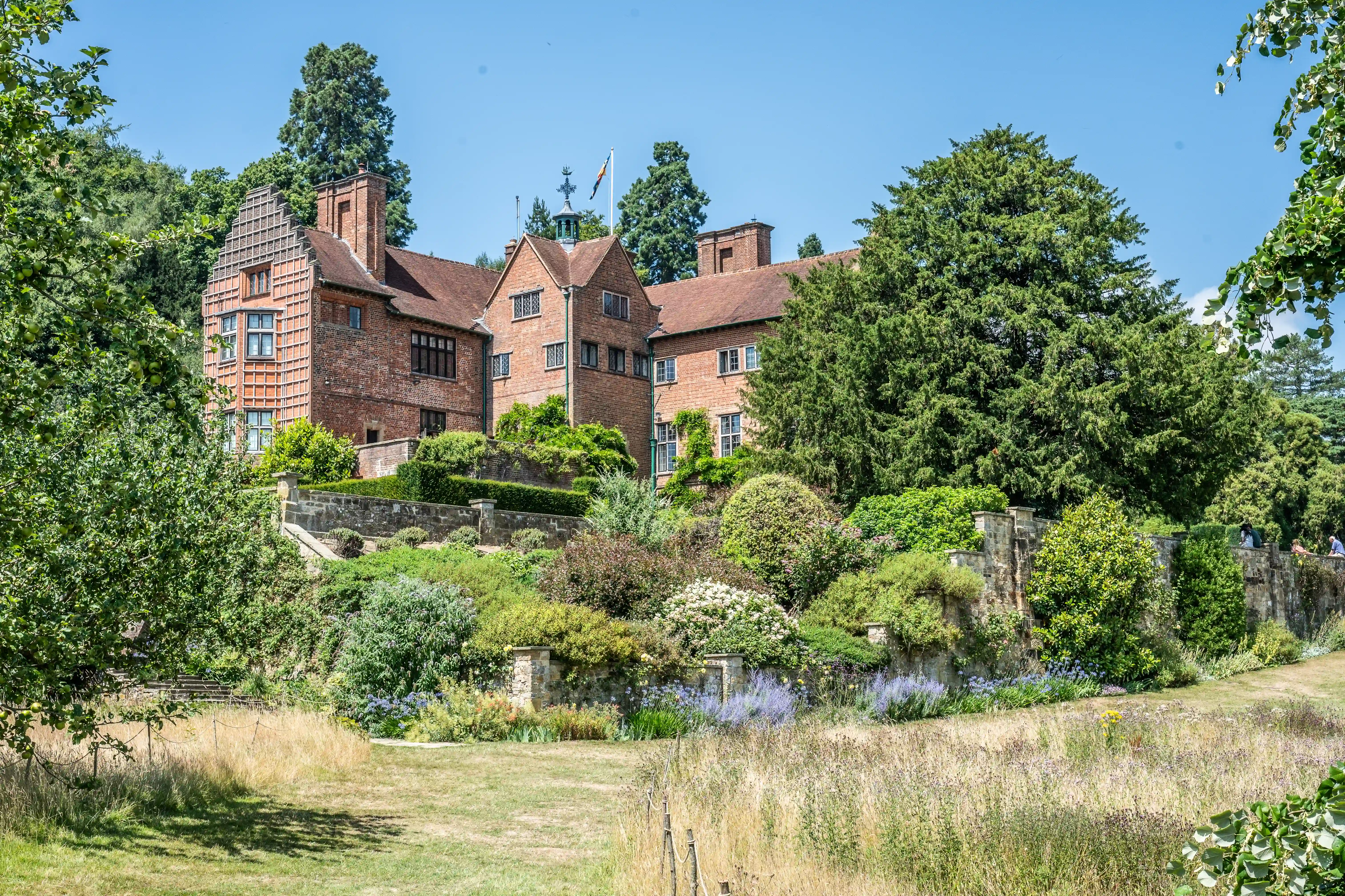 The image depicts a large, picturesque house made of red brick with a well-maintained garden in the foreground. The house features a mix of architectural styles, including a prominent tower-like structure on the left side. The garden is lush with various plants, flowers, and neatly trimmed hedges. The scene is set on a bright, sunny day with a clear blue sky, and the house is surrounded by tall trees, adding to the serene and idyllic atmosphere.