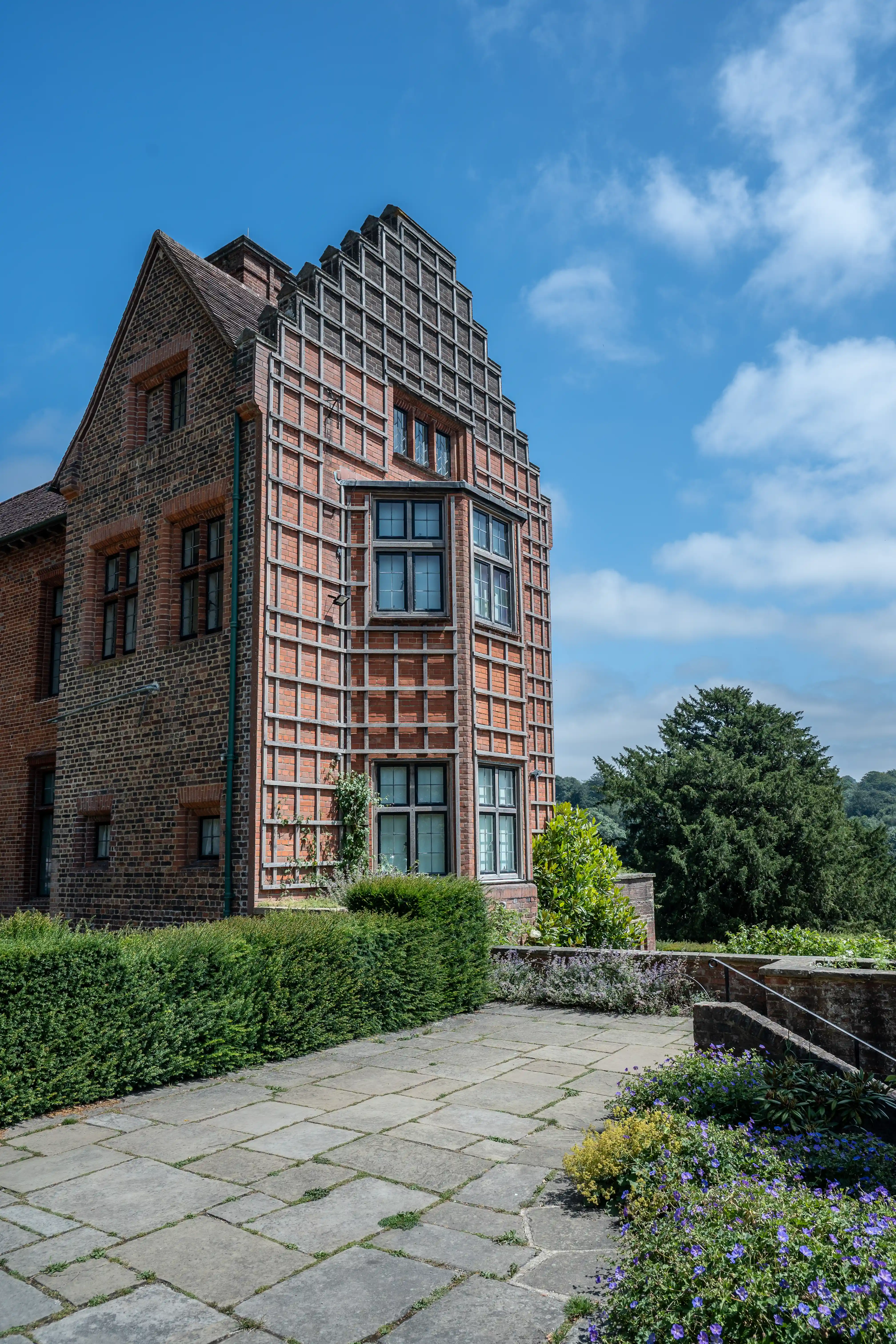 This image captures a charming brick manor house set against a backdrop of lush greenery and a bright blue sky dotted with fluffy clouds. In the foreground, a vibrant flowerbed overflows with delicate purple and white blooms, leading the eye towards the historic building. The manor itself boasts distinctive red brickwork, multiple chimneys, and traditional windows, hinting at a rich past. A flag flutters proudly atop one of the chimneys. The overall scene evokes a sense of tranquil, timeless English countryside living.