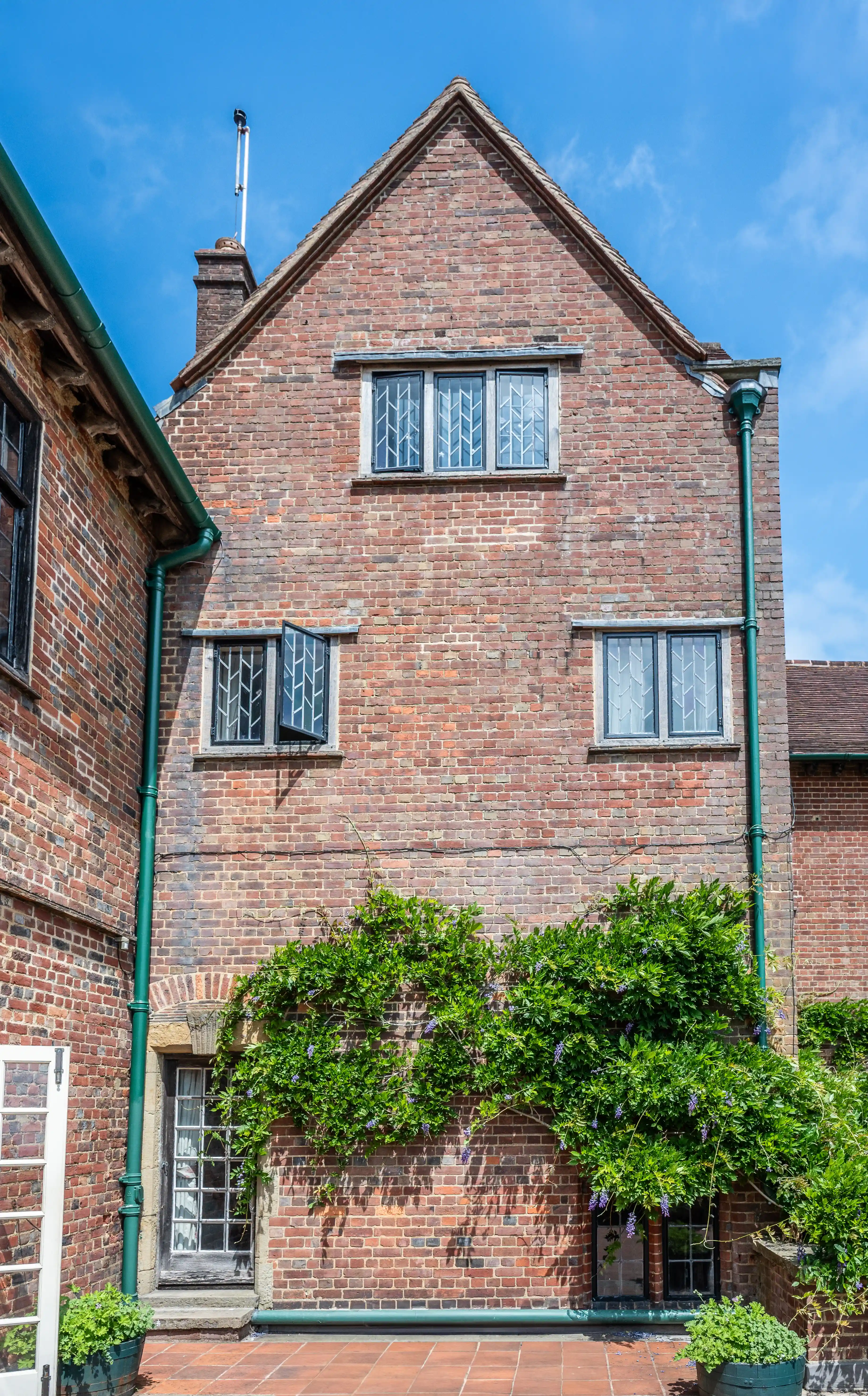 The image depicts a traditional brick building with a steep, triangular roof. The structure features multiple windows with lattice panes and green downspouts. The facade is adorned with lush green plants, and the ground level has a tiled patio area with potted plants. The building exudes a quaint, historical charm.