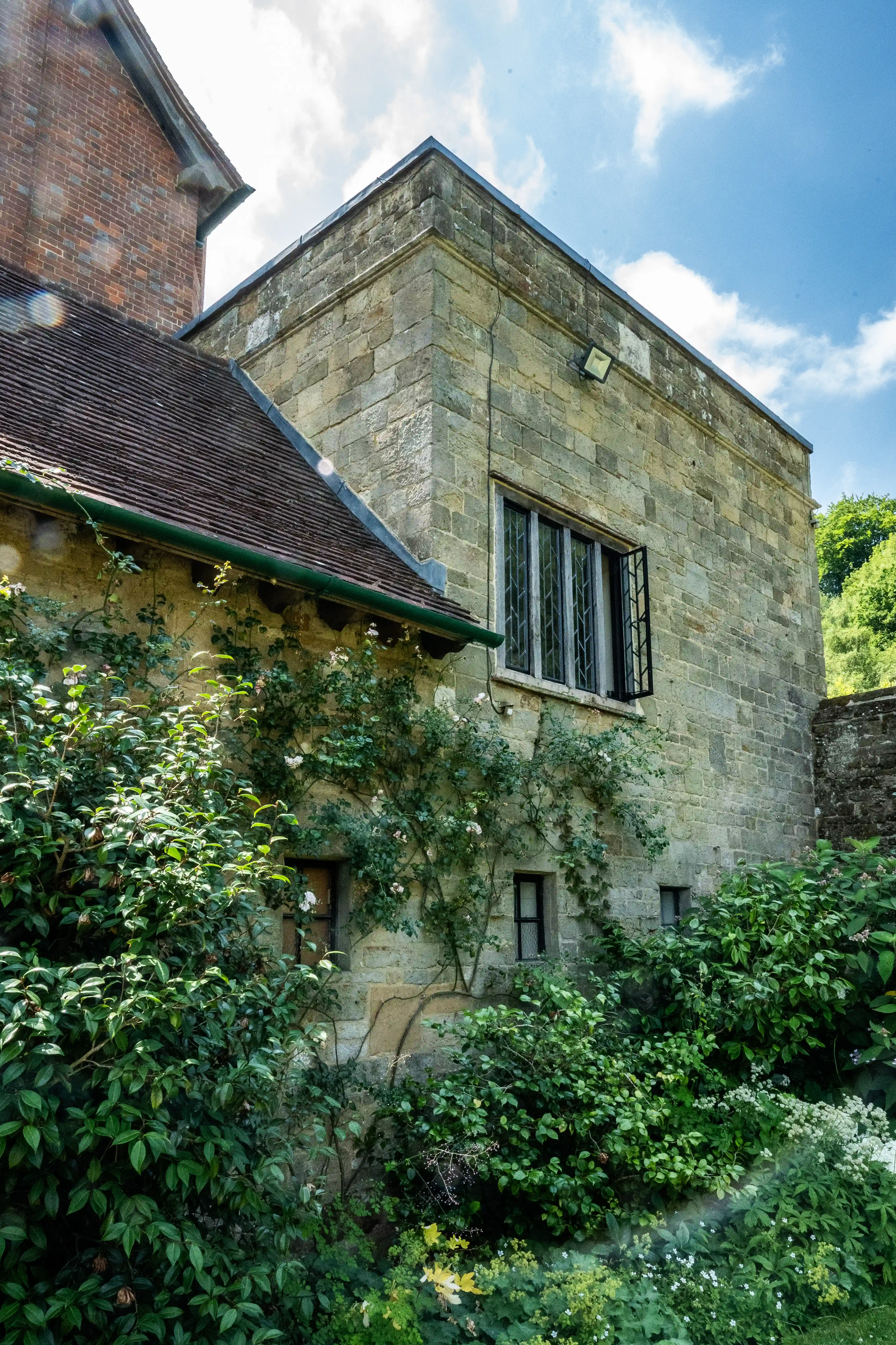The image depicts a rustic stone building with a tiled roof, partially covered by lush greenery and climbing plants. The structure features small, barred windows and is situated in a verdant garden. The building appears to be old, with visible wear and character, suggesting historical significance. The scene is set on a bright, sunny day with a clear blue sky.