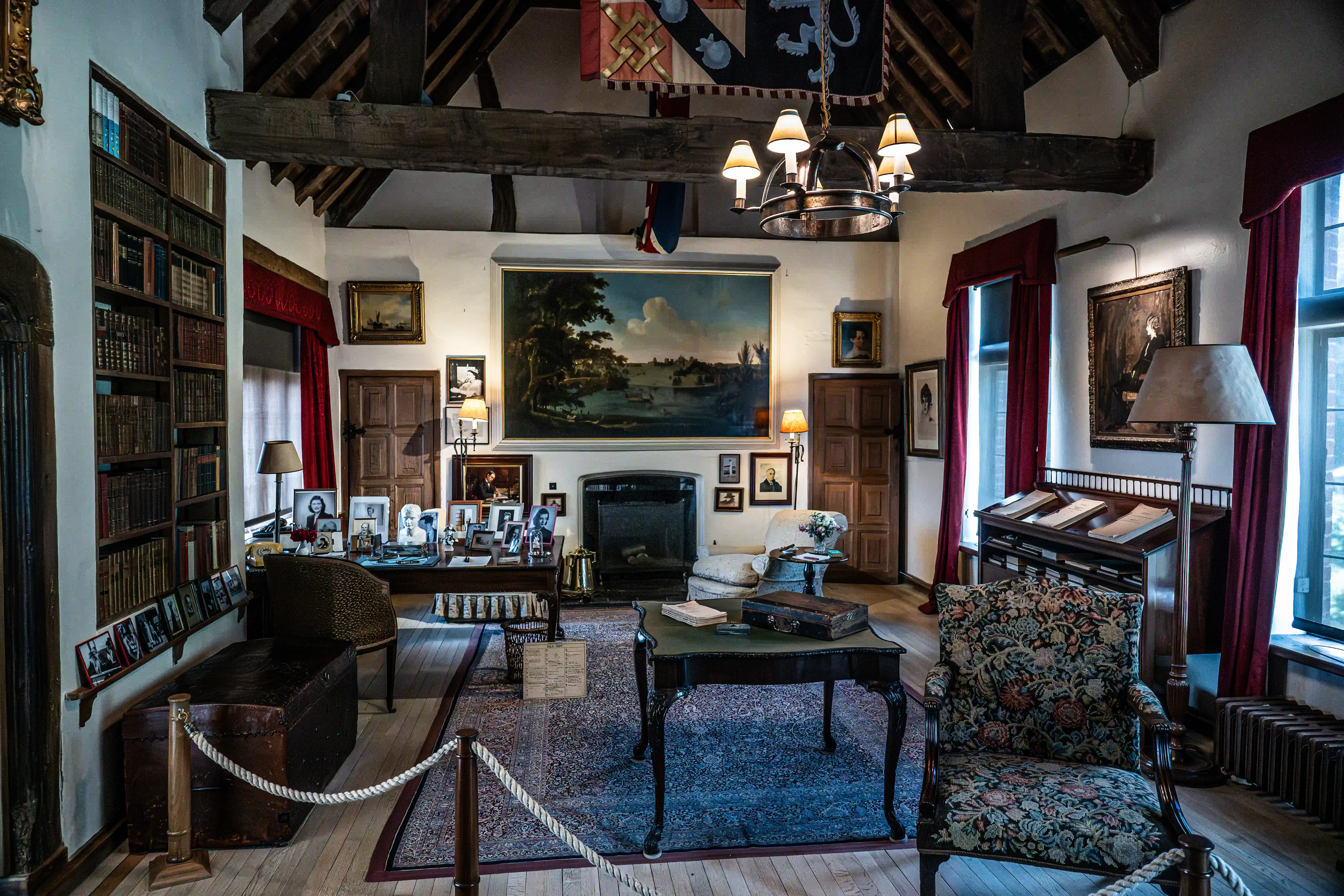 The image depicts a traditional, well-furnished study or library. The room features wooden beams, a large fireplace, and numerous bookshelves filled with books. There are various pieces of furniture, including chairs, a desk, and a small table. The walls are adorned with several paintings and photographs, and the room is illuminated by a chandelier and several lamps.