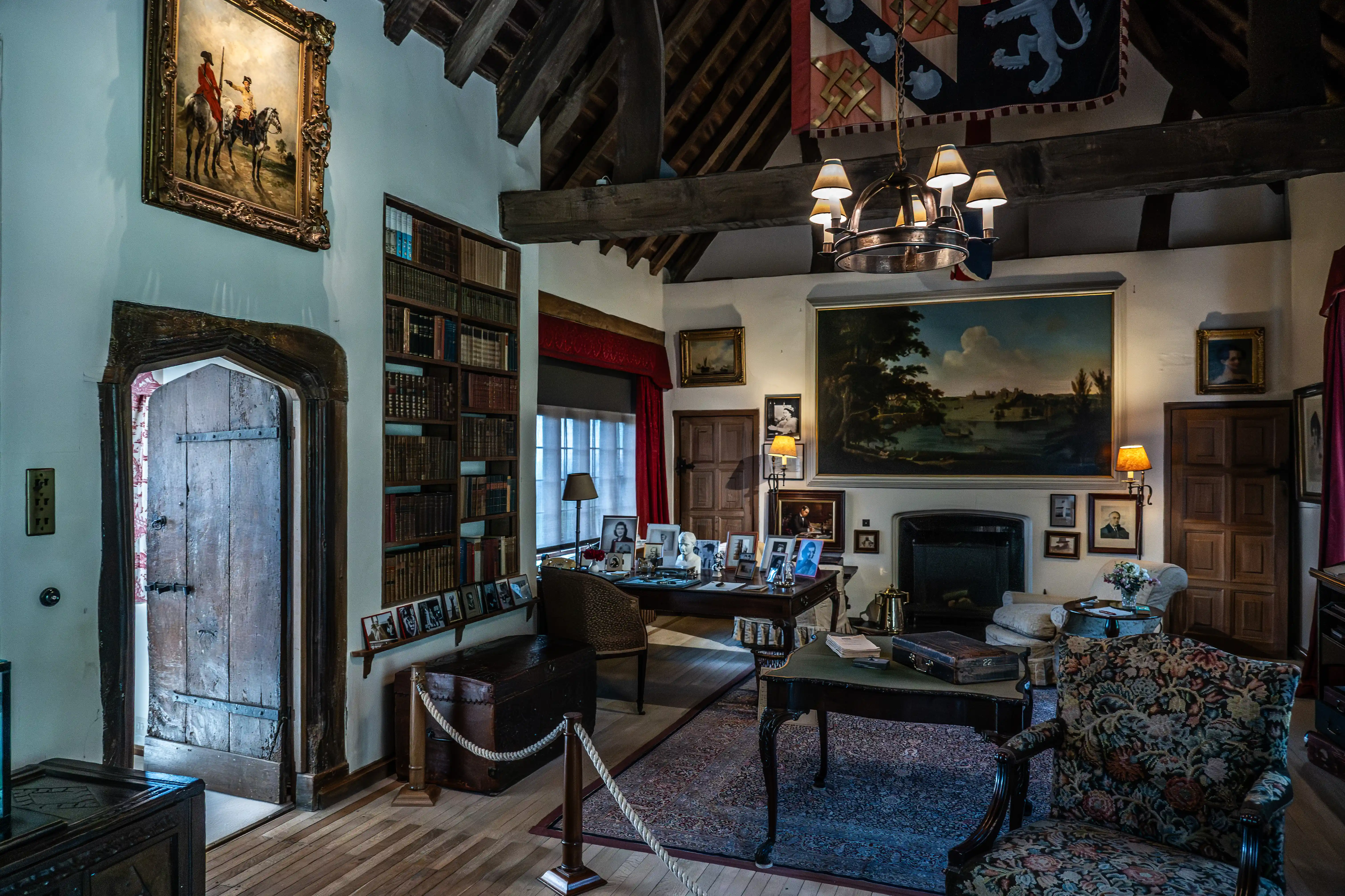 The image depicts a richly decorated room with a vintage and historical ambiance. The room features wooden beams on the ceiling, a large bookshelf filled with books, several framed paintings and photographs on the walls, and antique furniture including a desk, chairs, and a chest. A chandelier hangs from the ceiling, and there are multiple lamps providing light. The room also includes a fireplace and a large, ornate door, contributing to its classic and scholarly atmosphere.