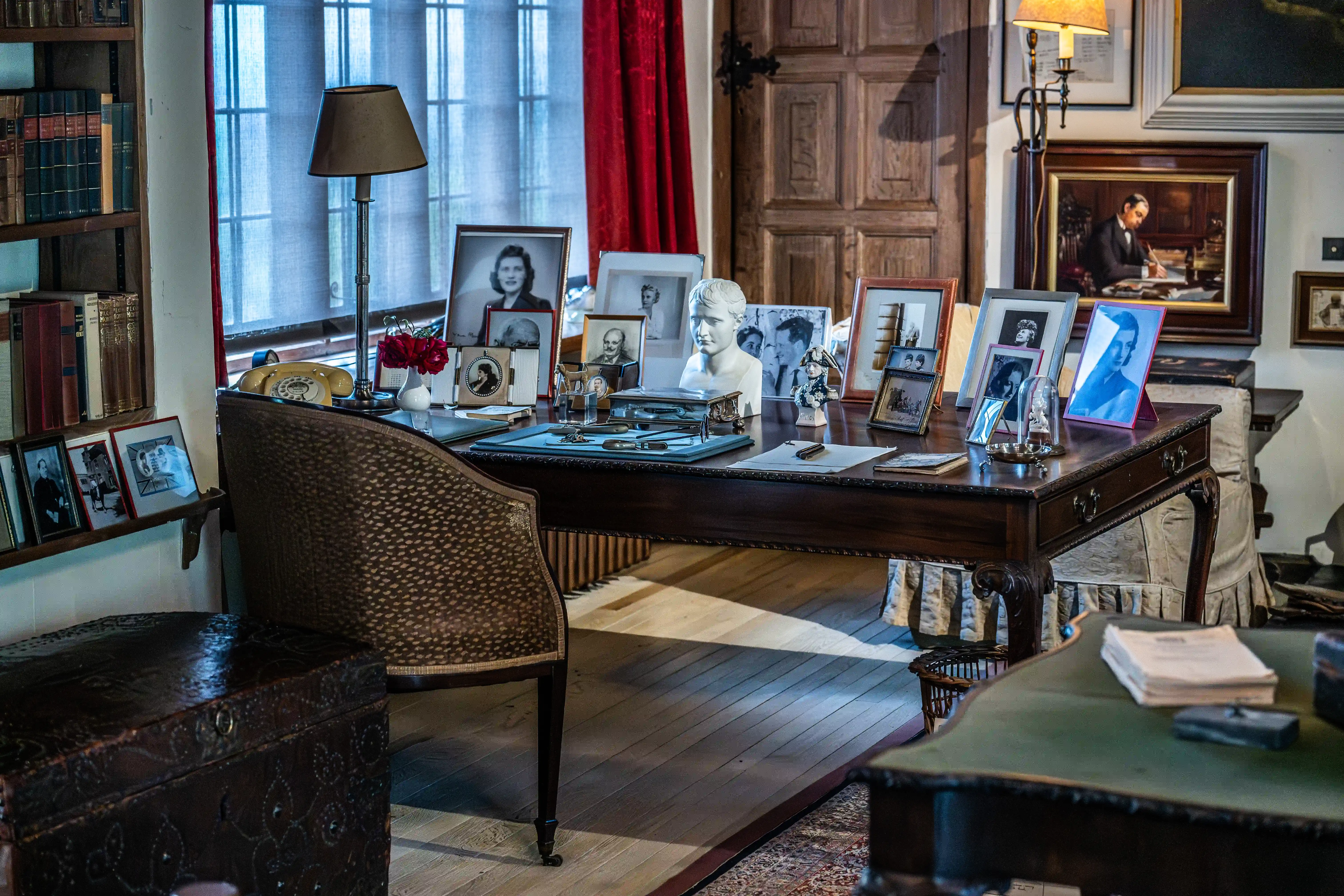 The image depicts a well-furnished study or office room with a wooden desk covered in framed photographs, a bust sculpture, and various personal items. The room also features a vintage telephone, a lamp, and a comfortable chair. There are bookshelves filled with books, and a large window with red curtains allowing natural light to enter. The walls are adorned with framed pictures and a painting, contributing to a cozy and scholarly atmosphere.