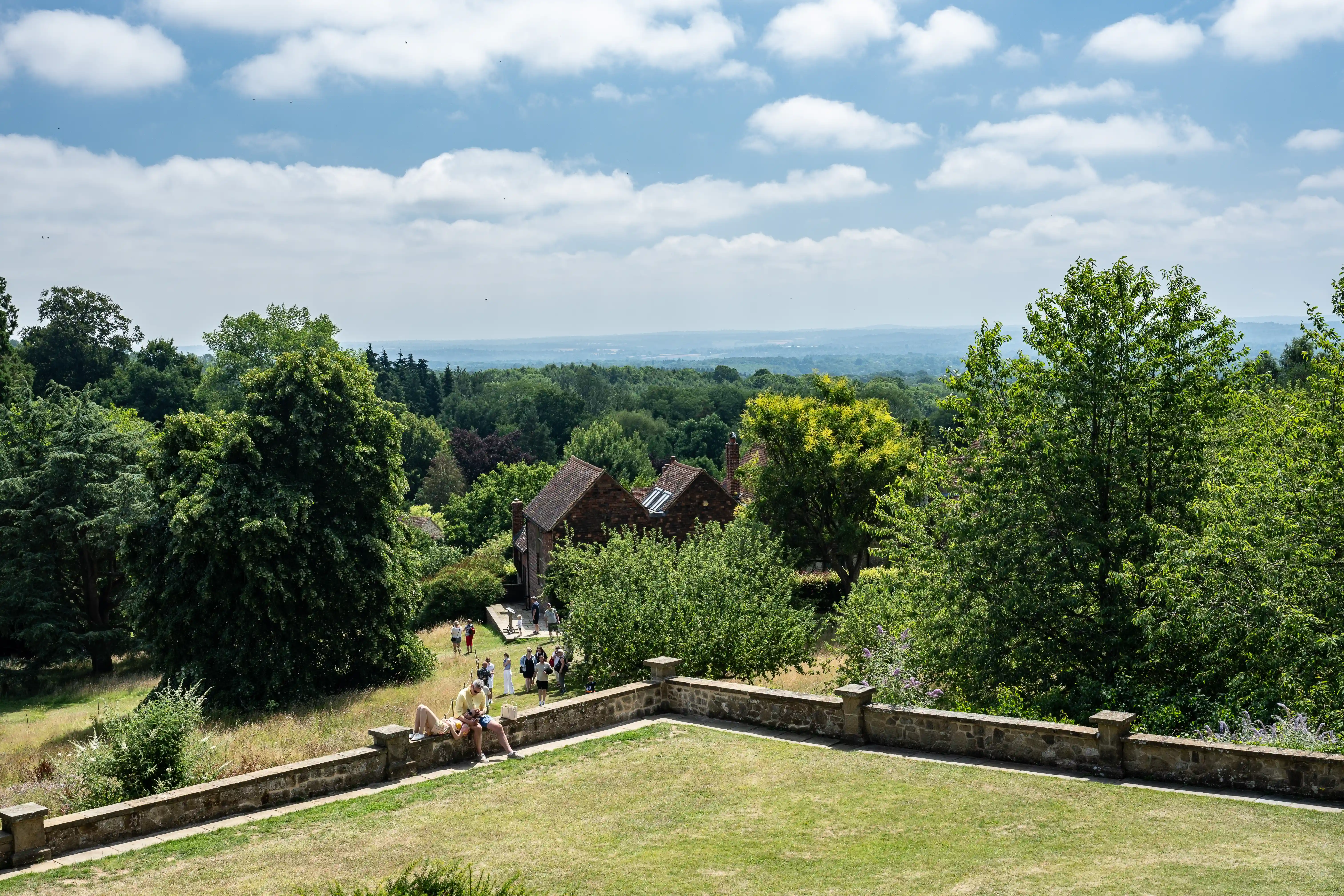 The image depicts a scenic view of a lush, green landscape with a variety of trees and vegetation. In the foreground, there is a stone terrace with a few people sitting and walking. Beyond the terrace, a group of people can be seen walking towards a rustic building with a red-tiled roof. The background reveals a vast expanse of forested area under a partly cloudy sky, extending to the horizon.