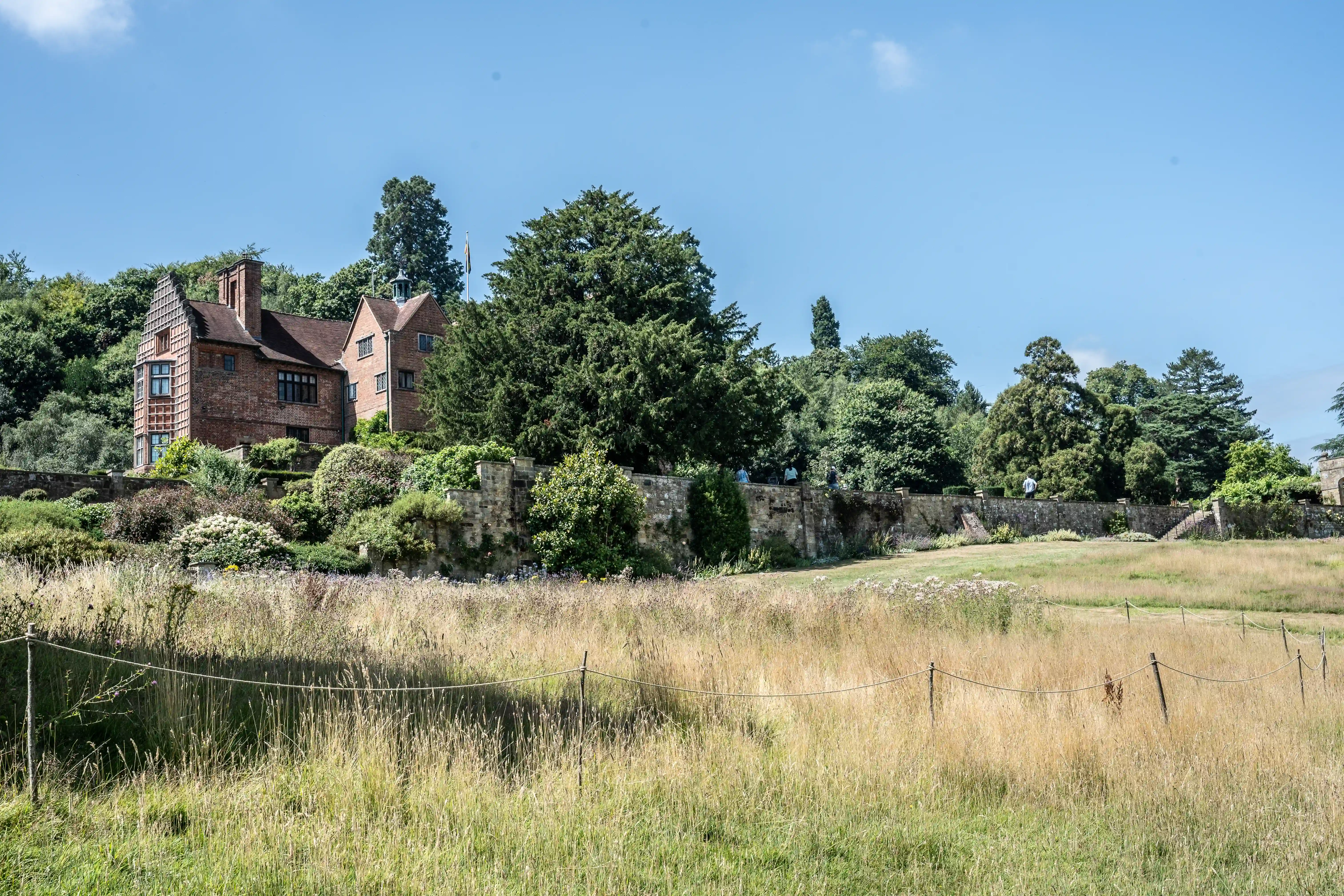 The image depicts a large, picturesque house with a brick exterior, surrounded by lush greenery and mature trees. The house is situated on elevated ground, with a stone wall running along the front. The foreground features a meadow with tall grasses and wildflowers, enclosed by a simple rope fence. The scene is set under a clear blue sky, giving the impression of a serene and well-maintained countryside estate.