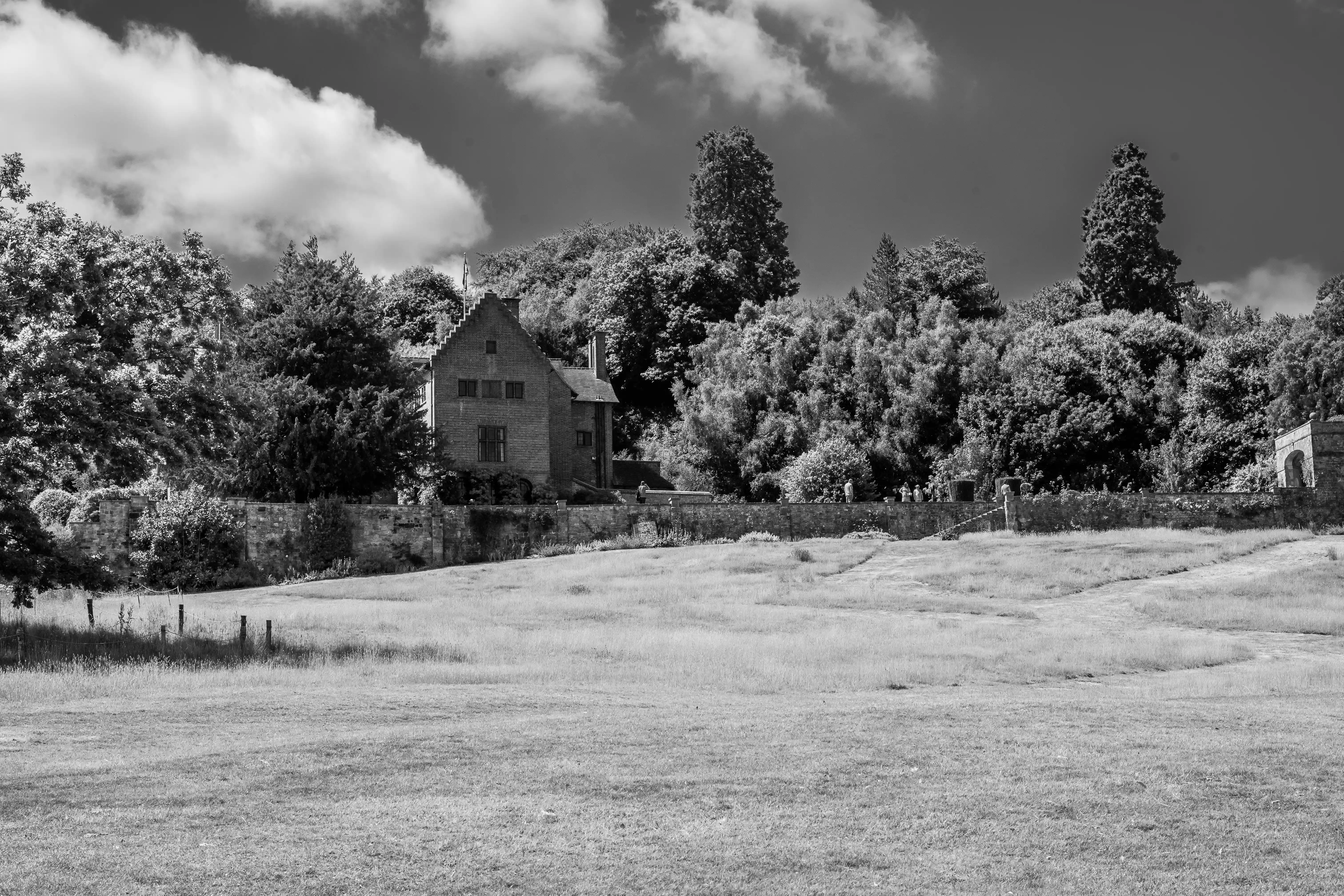 The image depicts a black and white photograph of a large, open grassy field with a few paths visible. In the background, there is a stone house surrounded by dense trees and foliage. The house has a traditional architectural style and is situated behind a stone wall. The sky above is partly cloudy, adding a dramatic effect to the scene.