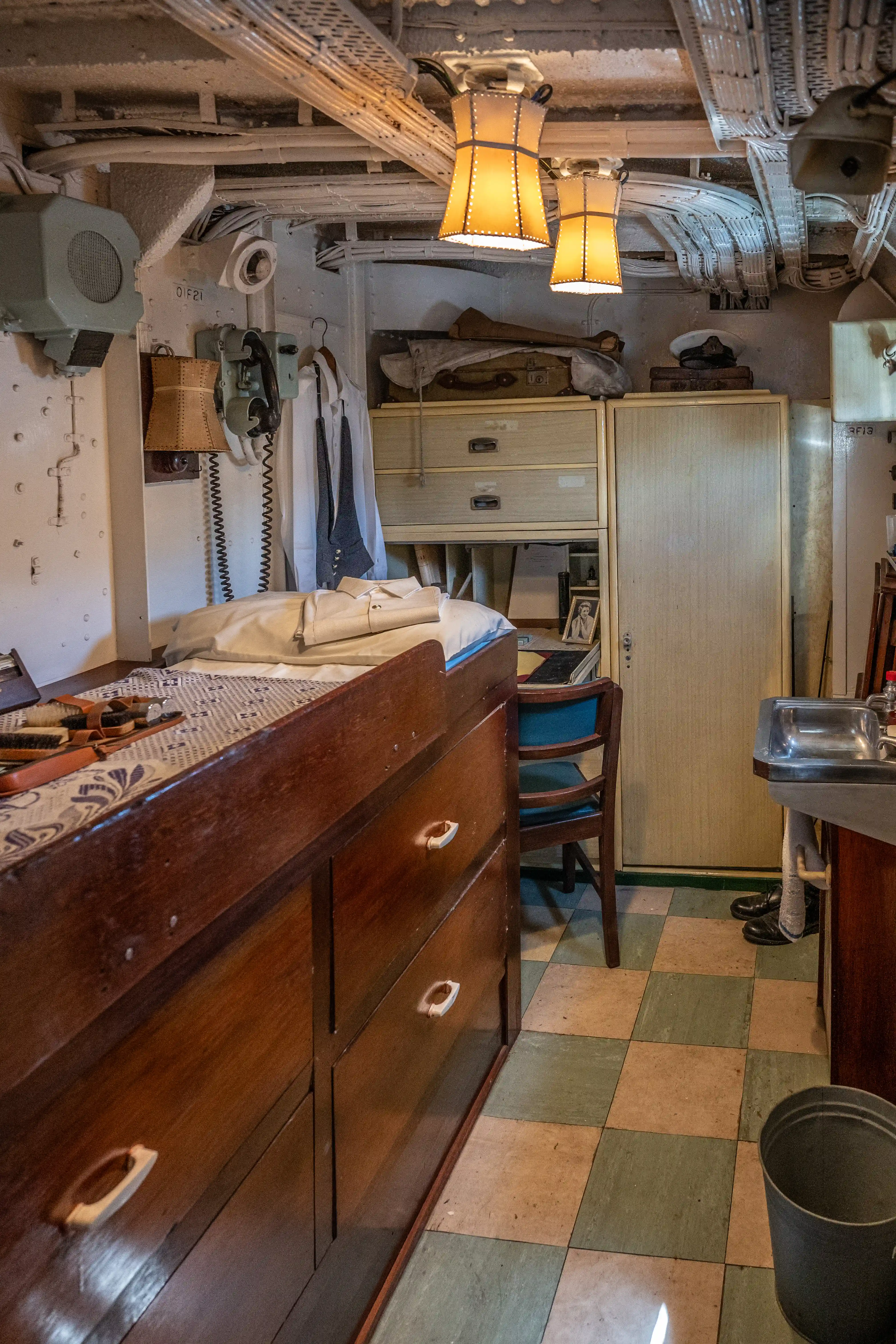 The image depicts a compact, utilitarian room that appears to be a cabin or small living quarters, possibly on a ship or submarine. The room contains a bed with a neatly arranged cover, a wooden dresser with multiple drawers, a chair, and a small sink. Various personal items and equipment are visible, including a telephone mounted on the wall, hanging clothes, and storage compartments. The floor is tiled in a checkered pattern, and the ceiling features exposed pipes and ventilation systems, along with two hanging lamps providing illumination.