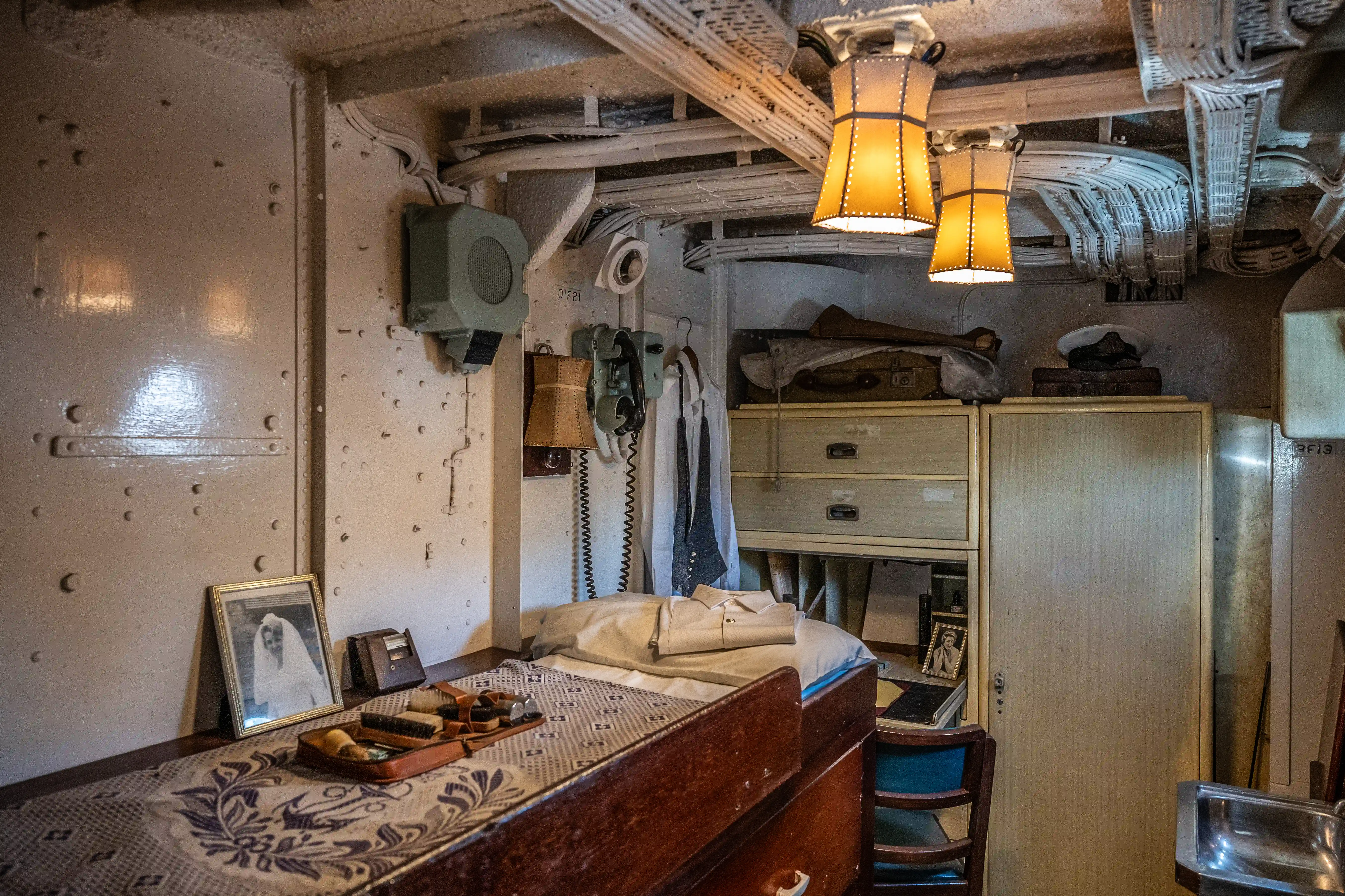 The image depicts a small, compact room that appears to be a cabin or quarters on a ship. The room contains a bed with a neatly folded shirt, a desk with personal items including a framed photograph, and various storage compartments. The walls are metallic with visible rivets, and there is a vintage telephone mounted on the wall. The lighting consists of two hanging lamps, and the overall decor suggests a historical or vintage setting.