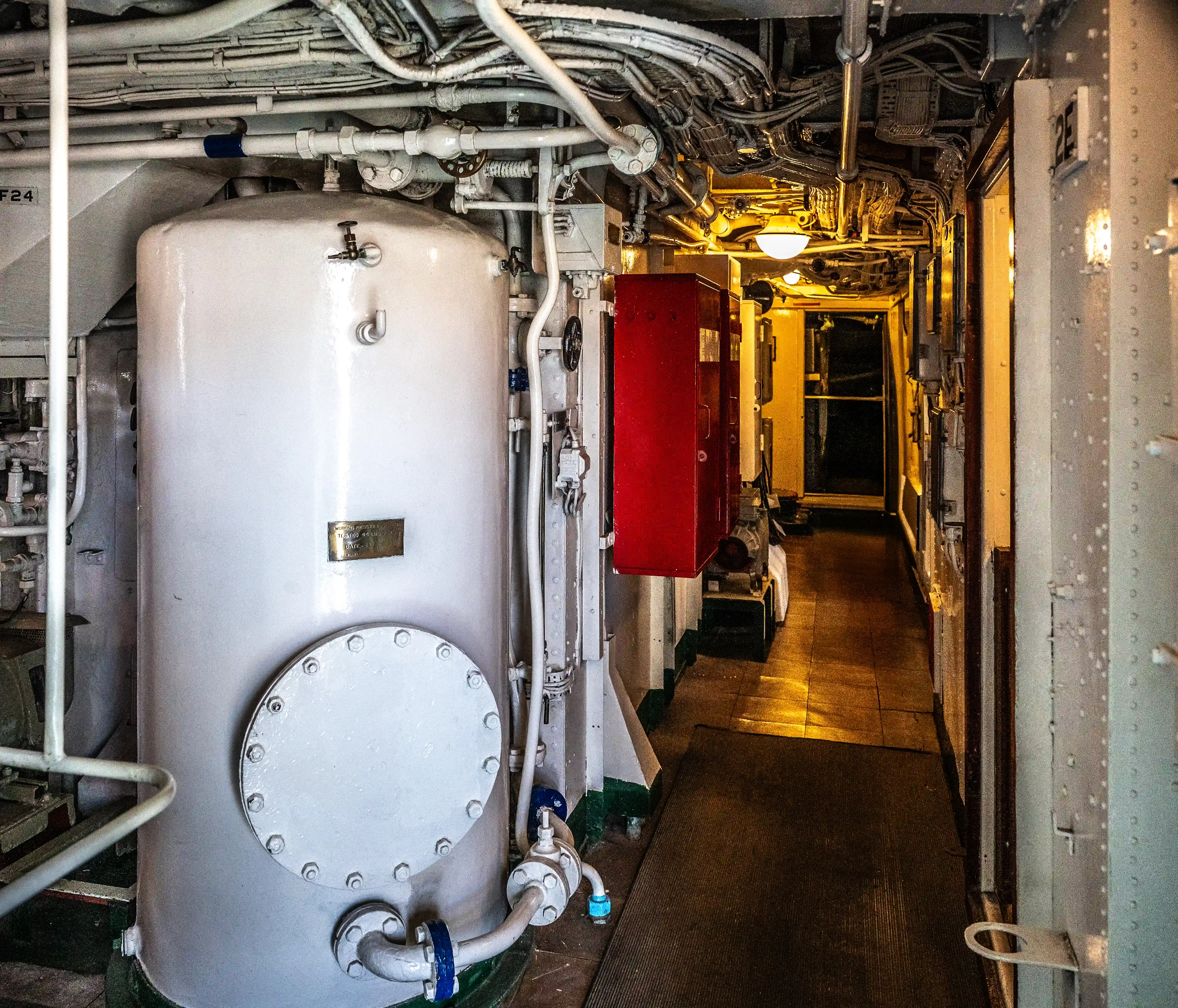 he image depicts an industrial or mechanical room, likely within a ship or submarine, characterized by a narrow corridor with various pipes and equipment. A large cylindrical tank with a circular hatch is prominently visible on the left side. The corridor is well-lit and extends into the background, with a red cabinet and additional machinery visible along the way. The setting suggests a functional space designed for operational and maintenance activities.