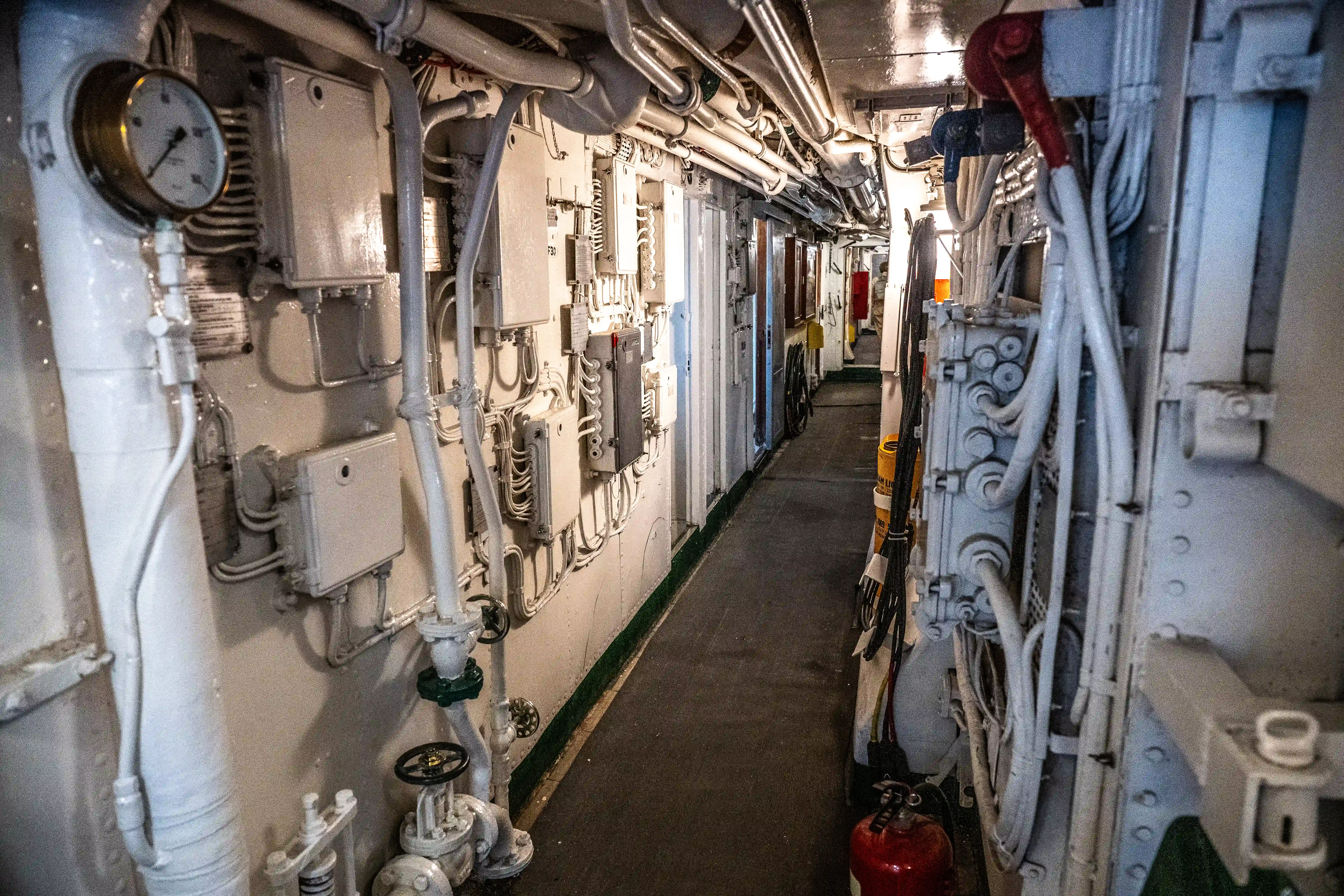 The image depicts a narrow, industrial corridor likely within a ship or submarine. The walls are lined with various mechanical components, pipes, and electrical panels. There is a pressure gauge mounted on the left wall, and a fire extinguisher is visible on the floor towards the end of the corridor. The space appears to be well-maintained and organized, with cables and equipment neatly arranged.