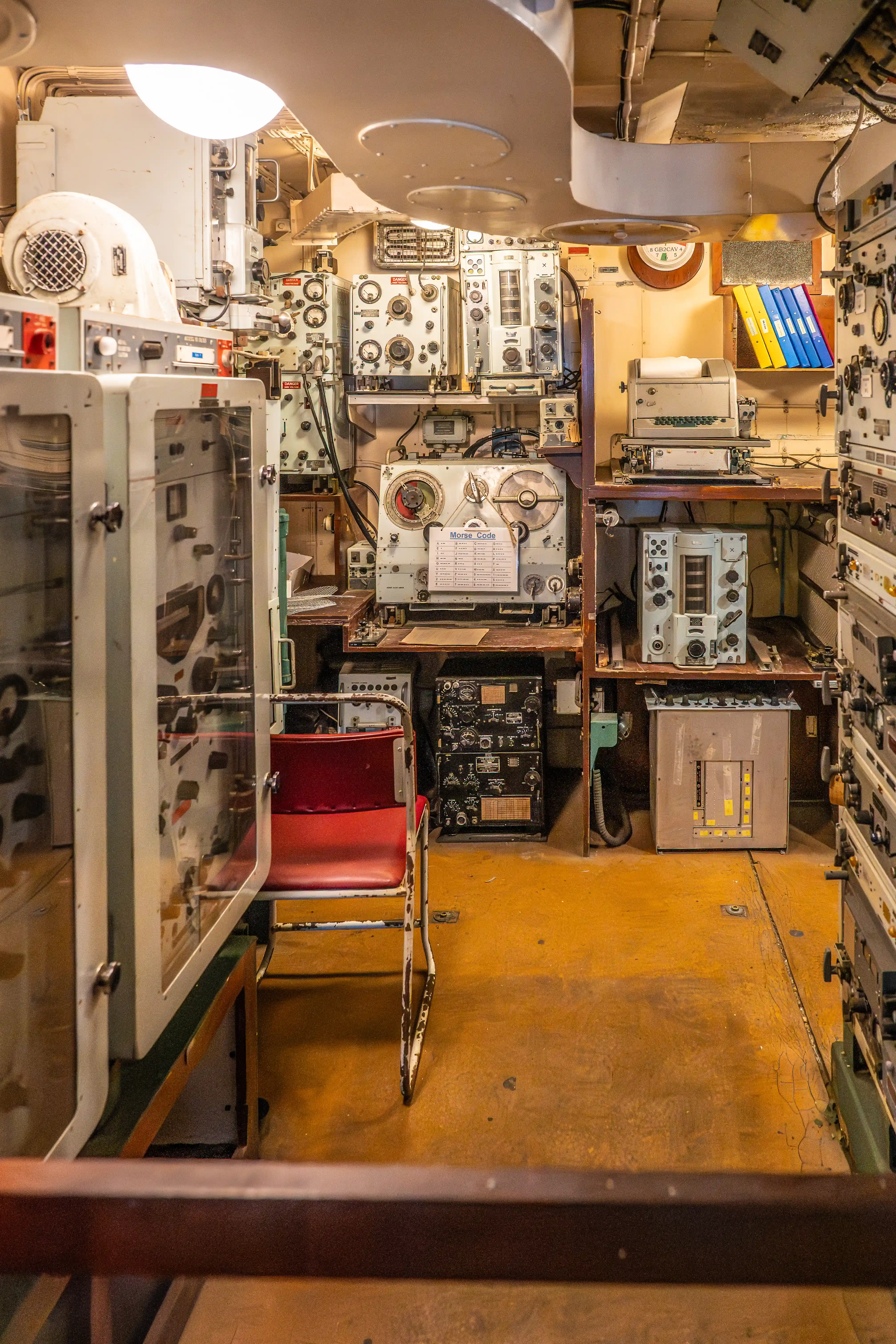 The image depicts the interior of a submarine control room, filled with various equipment and machinery. There are numerous control panels, communication devices, and other technical instruments. A red chair is visible in the center, and there are shelves with books and manuals. The space is compact and densely packed with equipment essential for submarine operations.