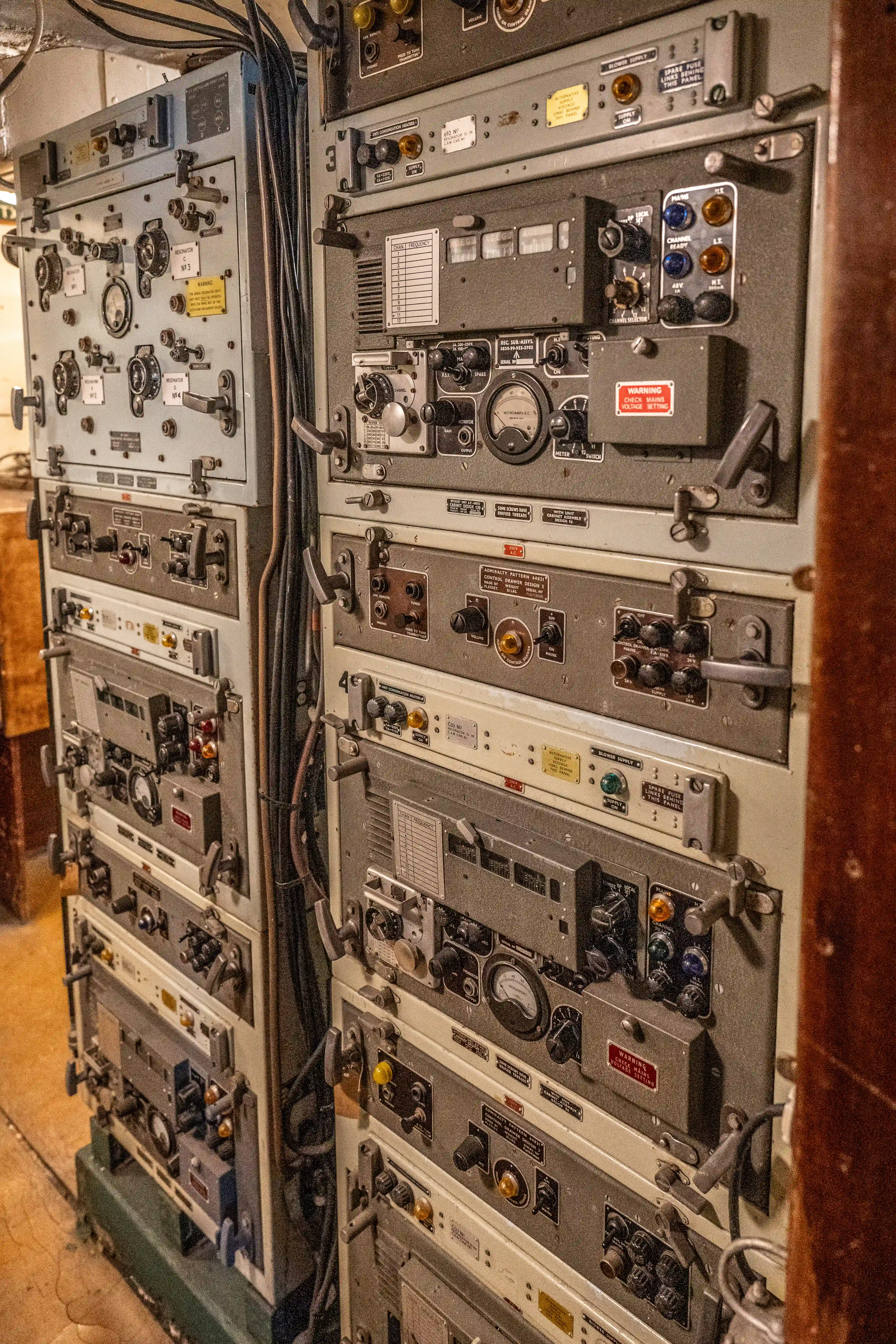 The image depicts a complex control panel with numerous switches, dials, and gauges. The equipment appears to be part of a vintage or industrial setup, possibly from a ship or a manufacturing plant. Various labels and warnings are visible on the panels, indicating specific operational instructions and cautions. The arrangement suggests a highly technical and specialized environment, likely used for monitoring and controlling machinery or systems.