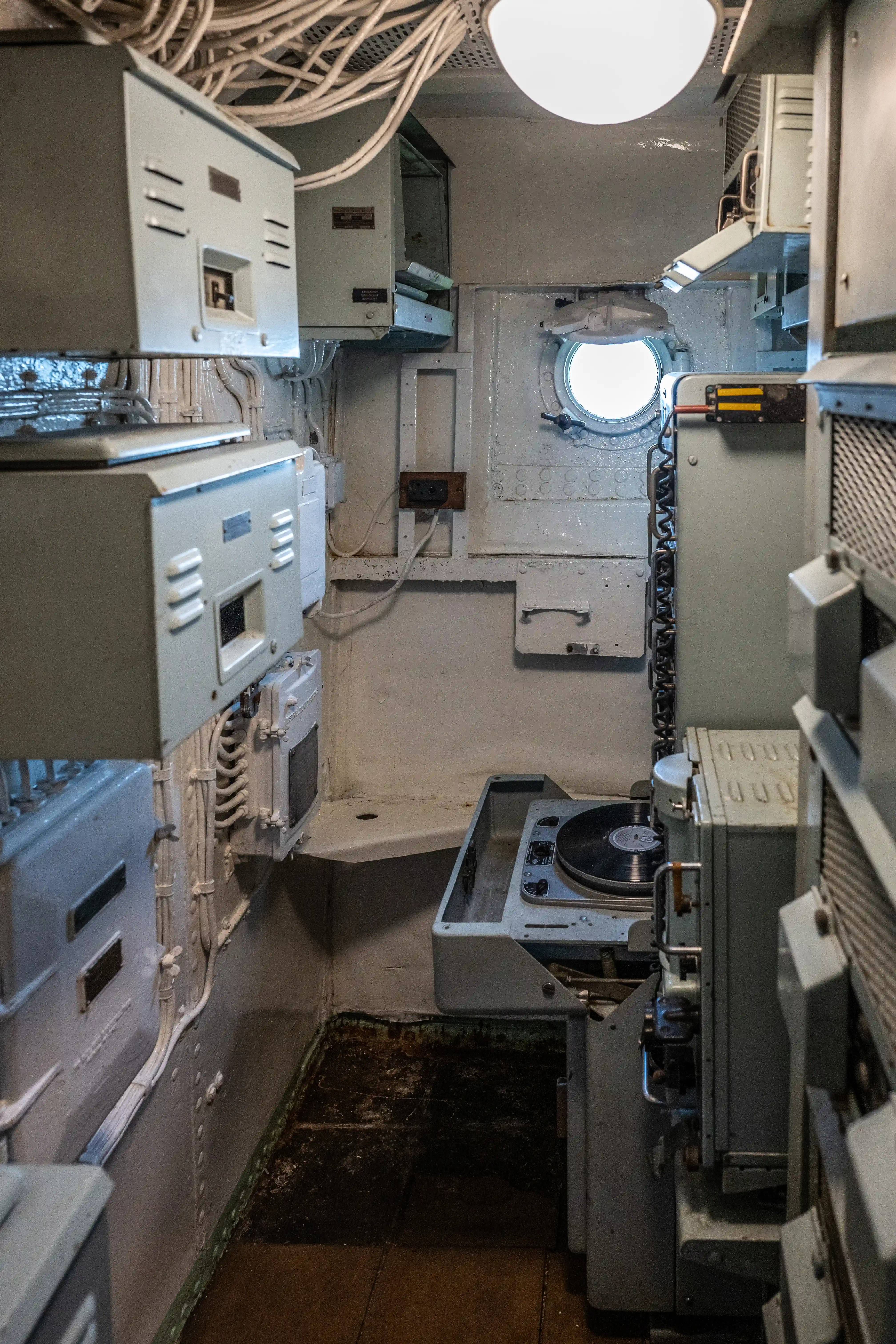 The image depicts the interior of a confined, industrial space, likely a control room or compartment within a submarine or ship. The area is filled with various equipment, control panels, and machinery, including a periscope or viewing port. The environment is characterized by a utilitarian design with a focus on functionality, featuring numerous cables, switches, and electronic devices. The lighting is provided by overhead fixtures, illuminating the workspace.