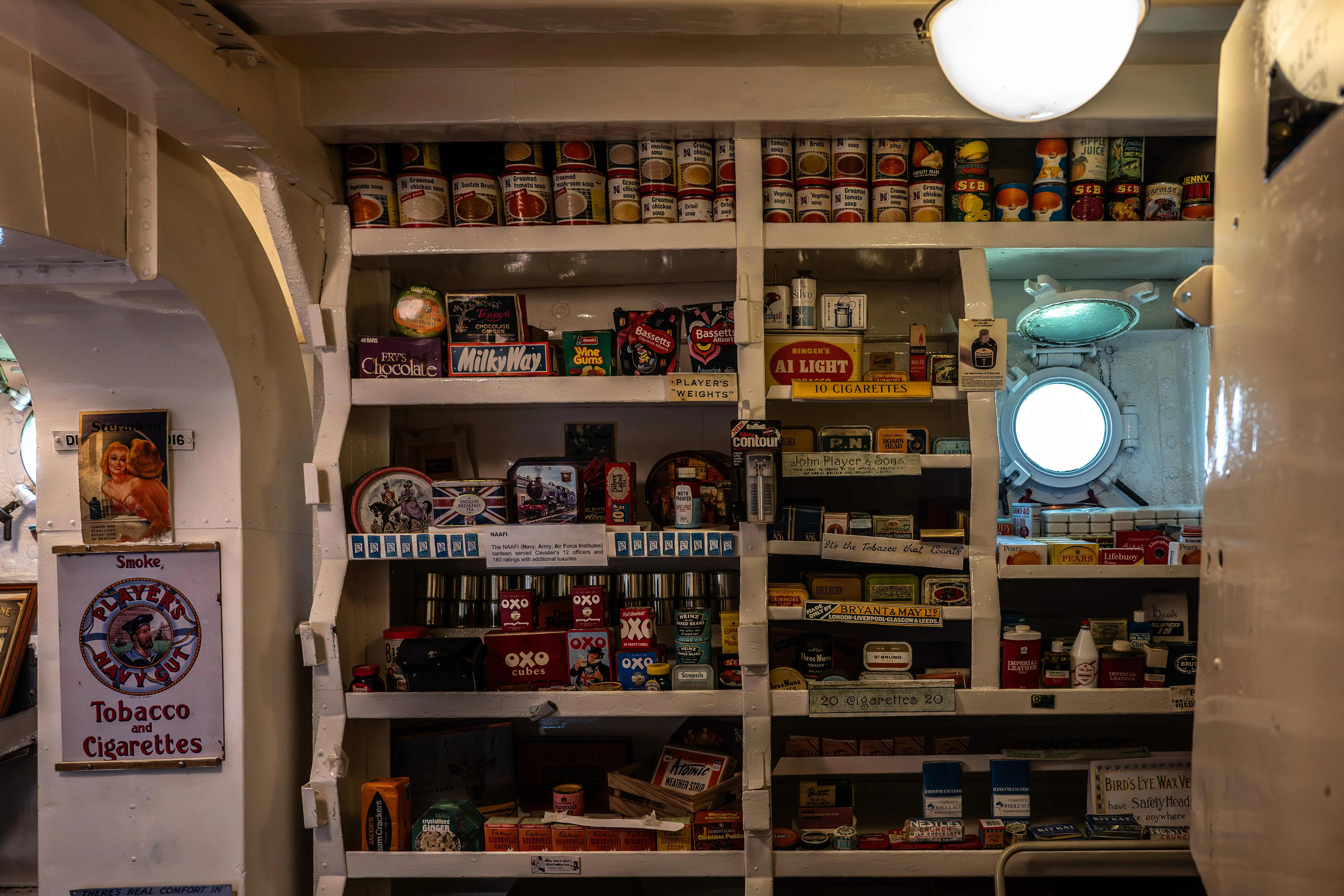 The image depicts a well-stocked ship's store or pantry with various vintage products. Shelves are filled with canned goods, tobacco products, and other supplies. Brands such as Player's Navy Cut, OXO, and Ringer's are visible. There is also a round porthole window on the right side, indicating the setting is on a ship. The overall scene suggests a historical or museum-like setting.