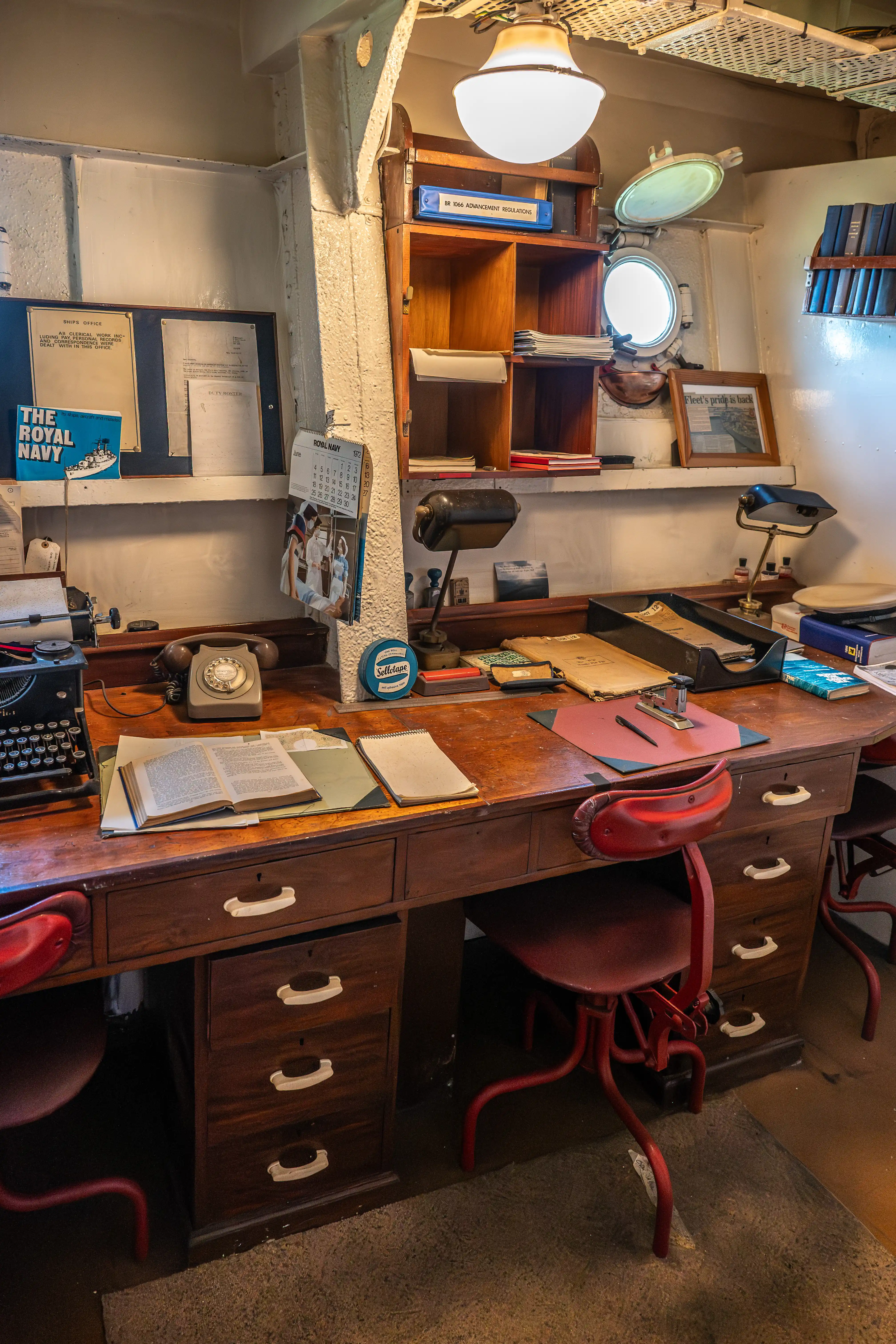 The image depicts a vintage office setting, likely from a naval ship, featuring a wooden desk with various items including an old-fashioned typewriter, a rotary phone, books, and documents. The desk is accompanied by a red chair and is situated in a small, well-lit room with a lamp, shelves, and various naval-related decorations and equipment. The space appears to be a historical recreation or museum exhibit, showcasing the working environment of a naval officer.