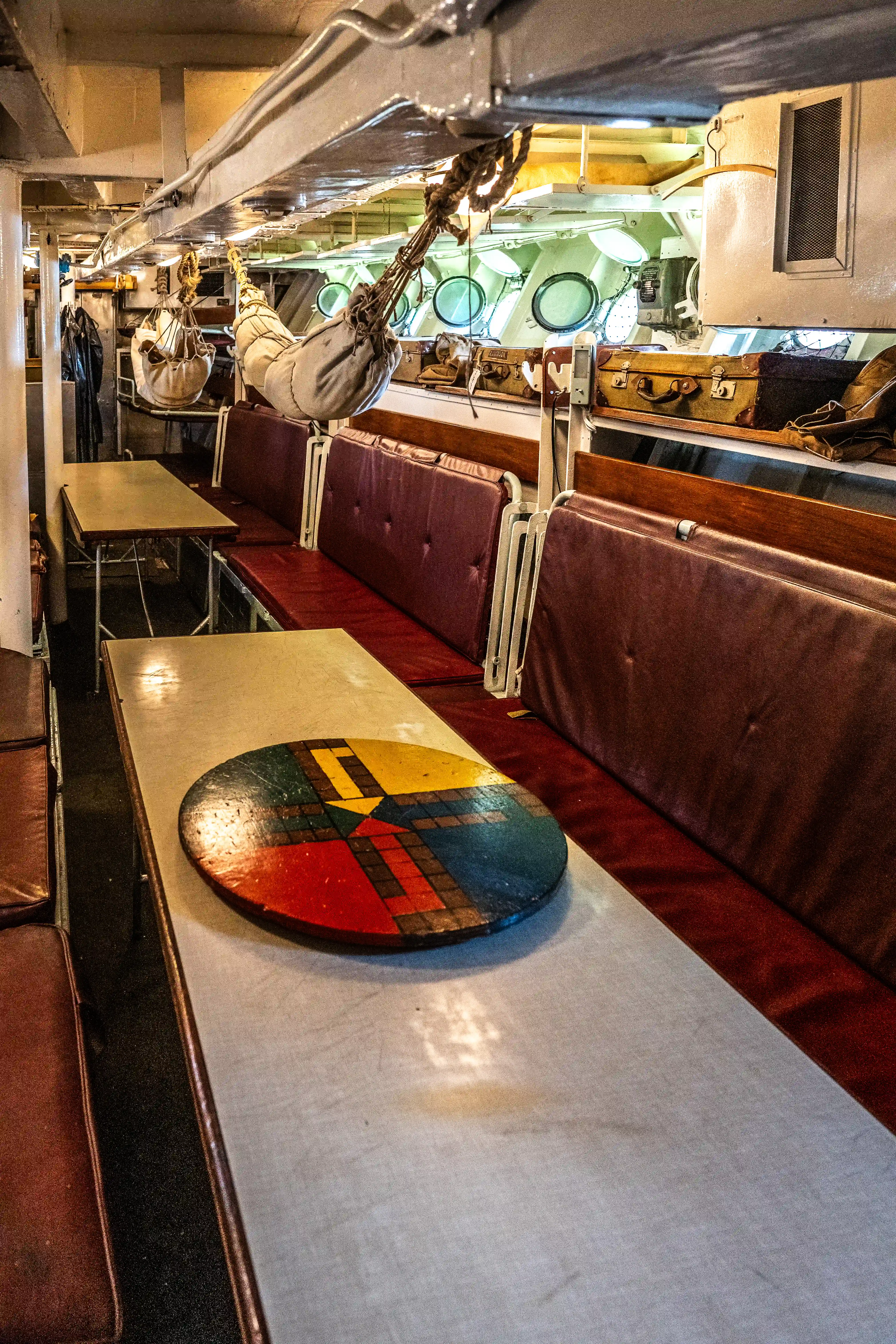 The image depicts the interior of a ship or submarine, showing a narrow corridor with seating booths on either side. The booths have red cushions and there is a table with a colorful, circular design in the foreground. Overhead, there are various items hanging, including bags and what appear to be hammocks. The background reveals a section with round windows, likely providing a view outside the vessel.