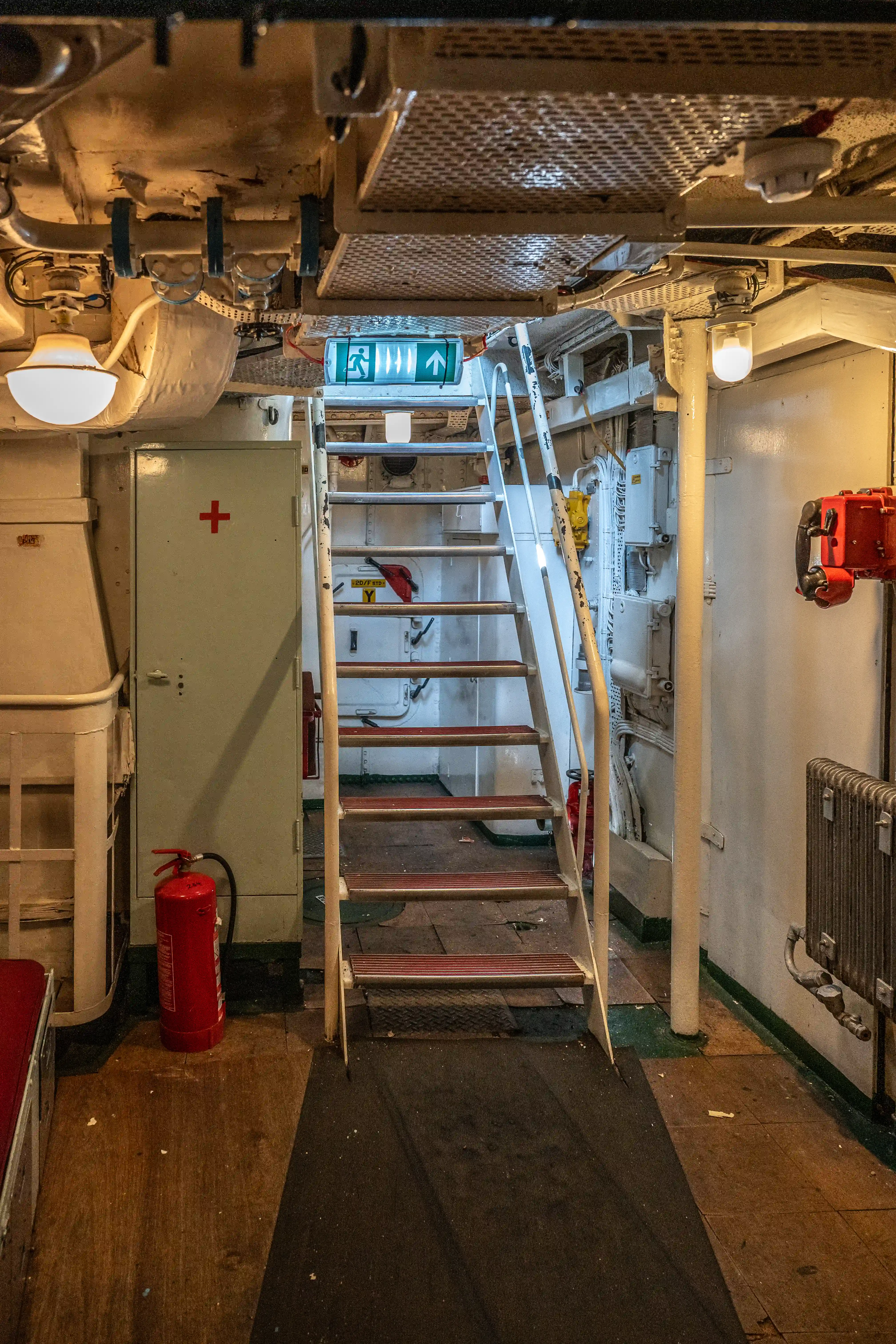 The image depicts an interior section of a ship or industrial facility. It shows a staircase leading to an upper level, with a green exit sign indicating the direction. There is a red fire extinguisher on the left side and various mechanical and electrical components visible. The area is well-lit with overhead lights and appears to be maintained for safety and functionality.