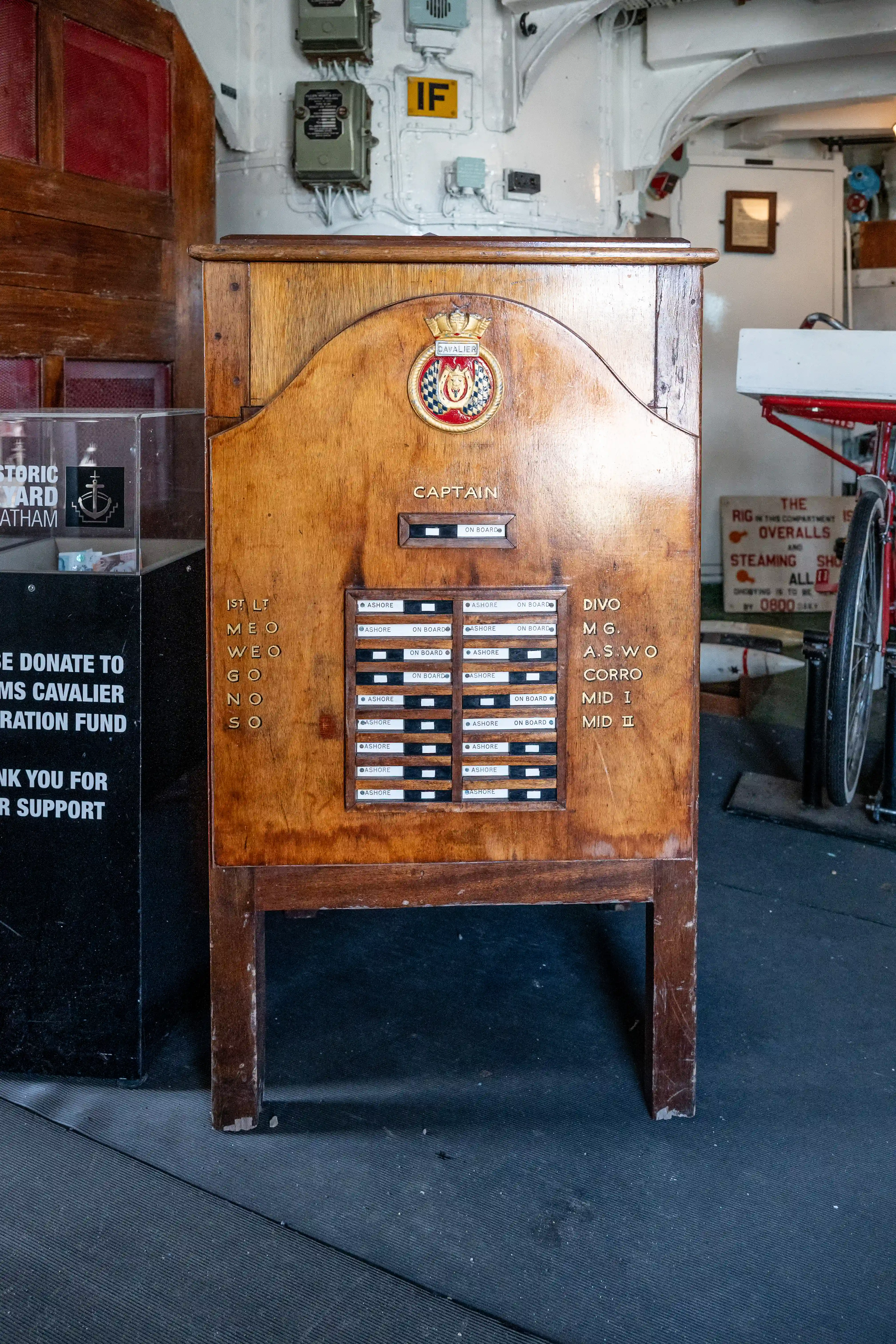 The image shows a wooden board used on a naval ship, likely for indicating the presence of personnel on board. The board has slots labeled with various ranks and positions such as Captain, 1st Lt, Divo, and others, with tags indicating 'Ashore' or 'On Board.' There is also a donation sign for a restoration fund and some naval equipment visible in the background.