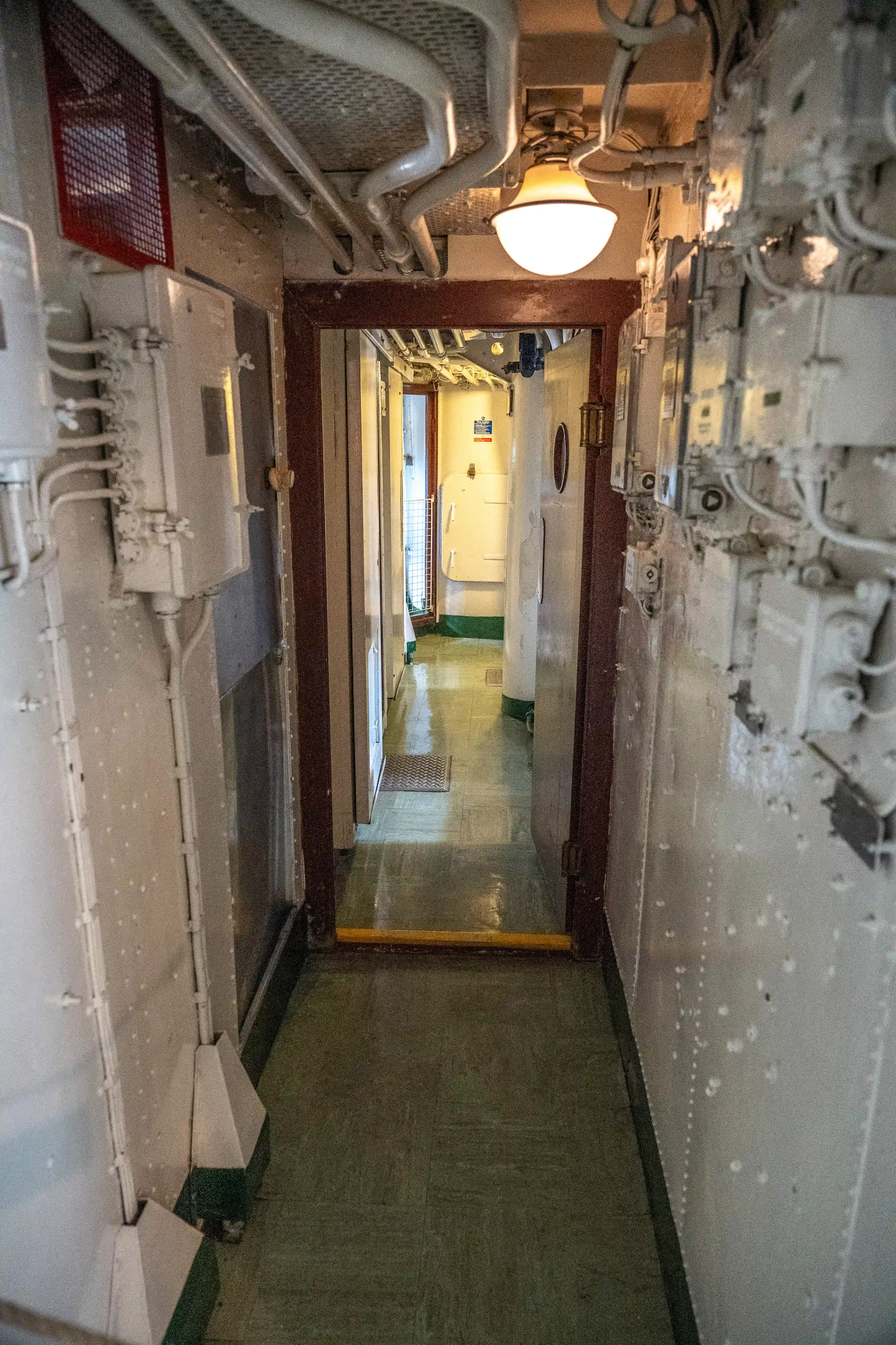 The image depicts a narrow, industrial-style hallway, likely within a ship or submarine. The corridor is lined with various mechanical and electrical components, pipes, and wiring. The floor is painted green, and the walls are white with visible bolts and fixtures. At the end of the hallway, there is a lit area with a door, suggesting further compartments or rooms beyond.