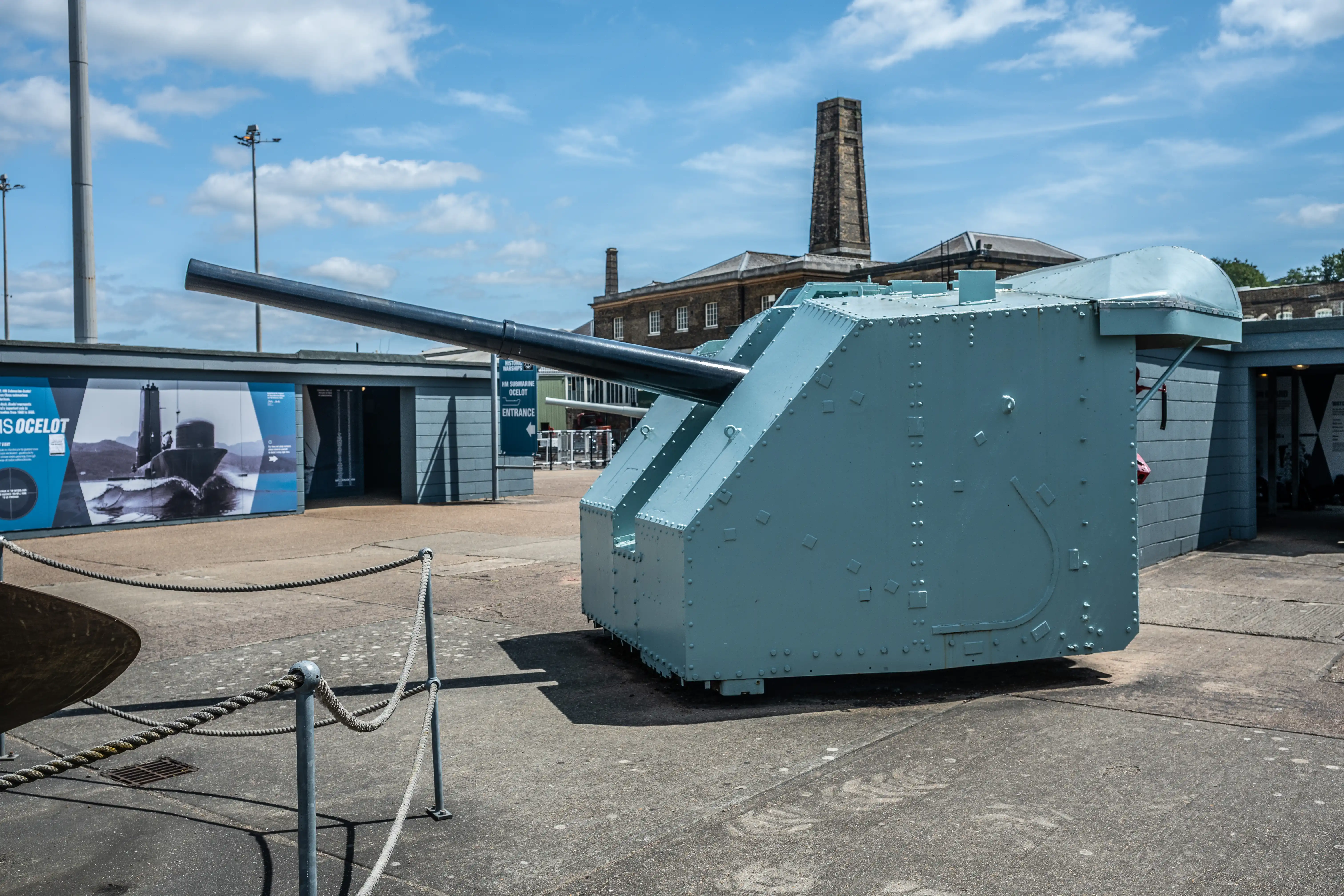 The image depicts a large naval gun turret, likely part of a historical warship or submarine, displayed outdoors in a museum setting. The turret is painted in a light blue-green color and is positioned prominently in the foreground. In the background, there is a building with a tall chimney and a sign indicating the entrance to an exhibit related to the HMS Ocelot, a submarine. The area is cordoned off with ropes, suggesting it is part of a protected exhibit.