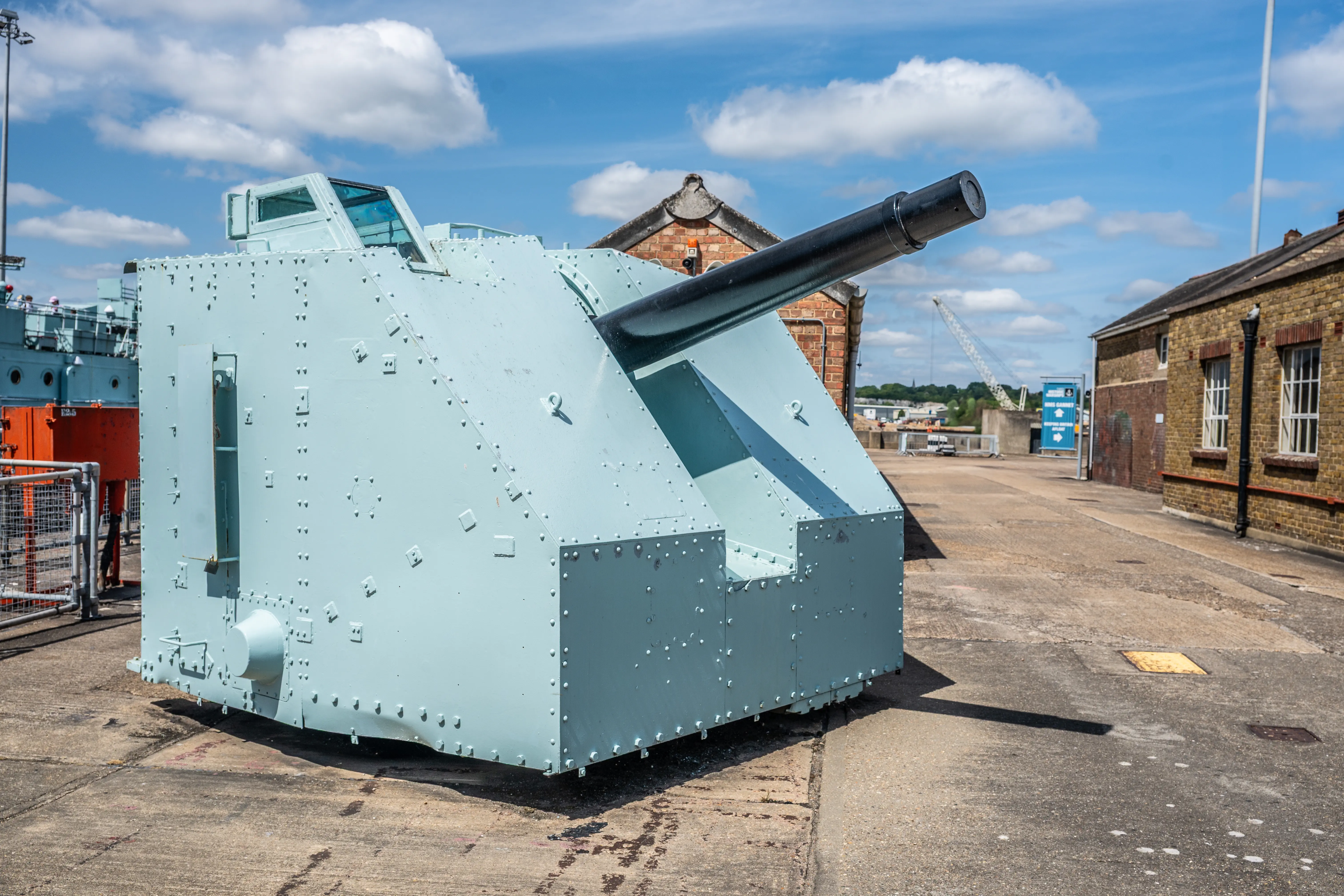 The image depicts an armored naval gun turret situated outdoors, likely in a historical or museum setting. The turret is painted in a light blue-green color and features a large cannon protruding from its angled front. The structure is riveted and has various fittings and reinforcements. In the background, there are brick buildings and a clear sky, suggesting a well-maintained area dedicated to preserving historical military artifacts.