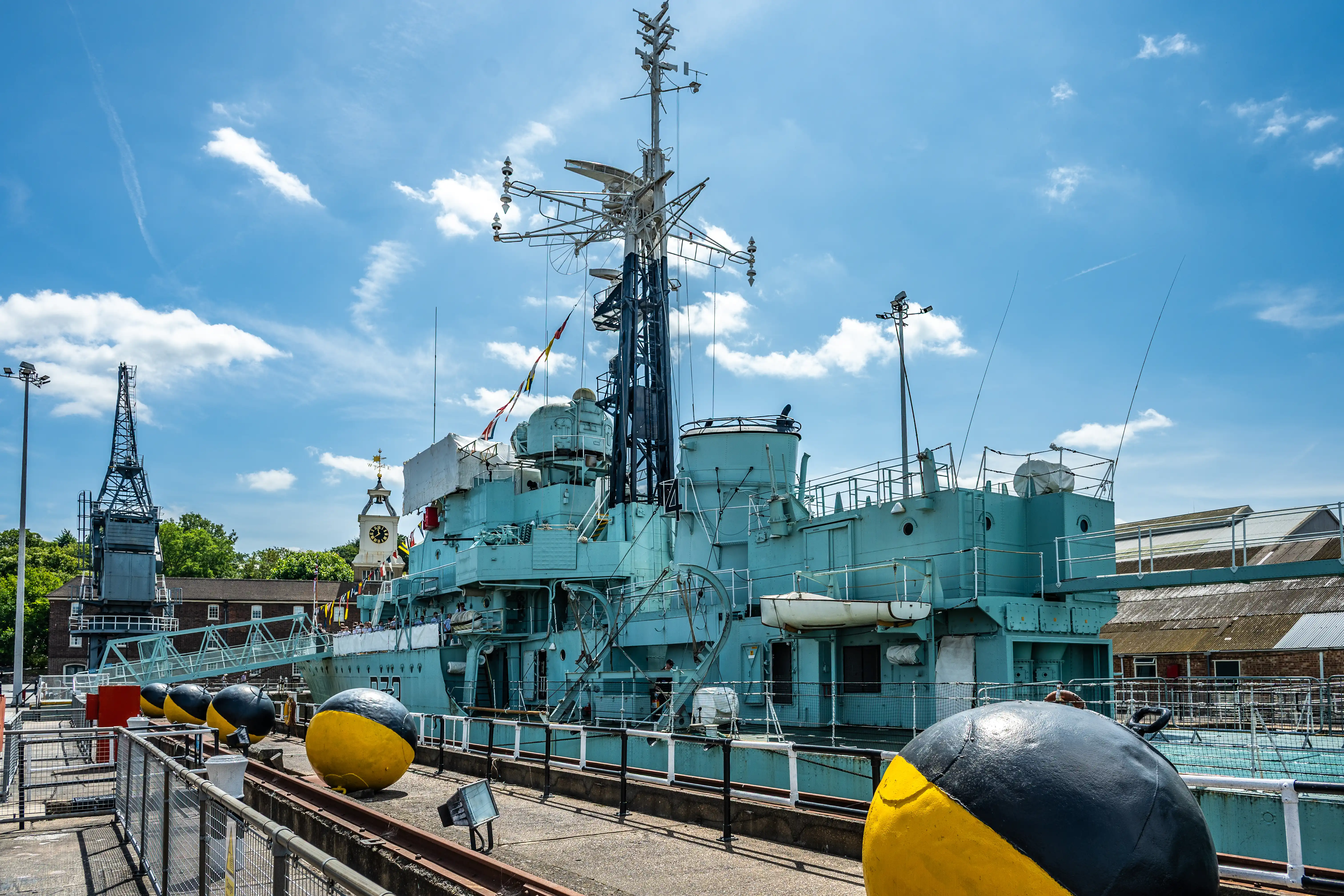 The image depicts a large naval ship docked in a port. The ship is painted in light blue and has various equipment and structures on its deck, including antennas and radar systems. The foreground features large spherical fenders with yellow and black markings. The background includes some buildings and a clock tower, under a bright, partly cloudy sky.